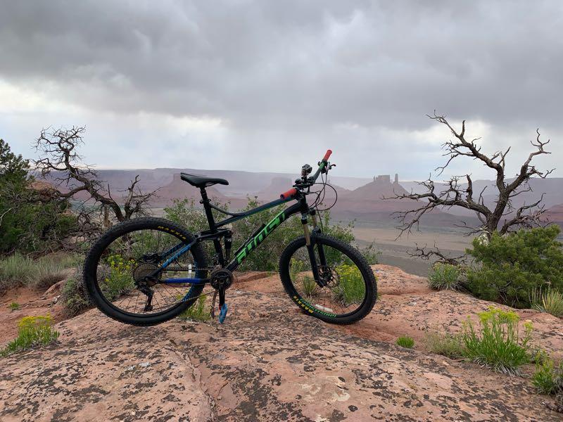 Mountain bike resting on rocky terrain with a scenic view of a valley and distant cliffs under a cloudy sky. Sparse vegetation and bare trees are visible, suggesting an outdoor adventure landscape. Porcupine Rim mountain bike trail.