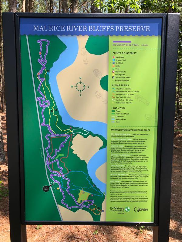 Map of Maurice River Bluffs Preserve, showing mountain bike and hiking trails, points of interest, land cover types, and rules for trail use. The map features a legend with different trail routes marked in various colors, including blue, orange, and yellow, with distances noted. North orientation is indicated, and the surrounding nature is visible in the background. The bluffs mountain bike trail.
