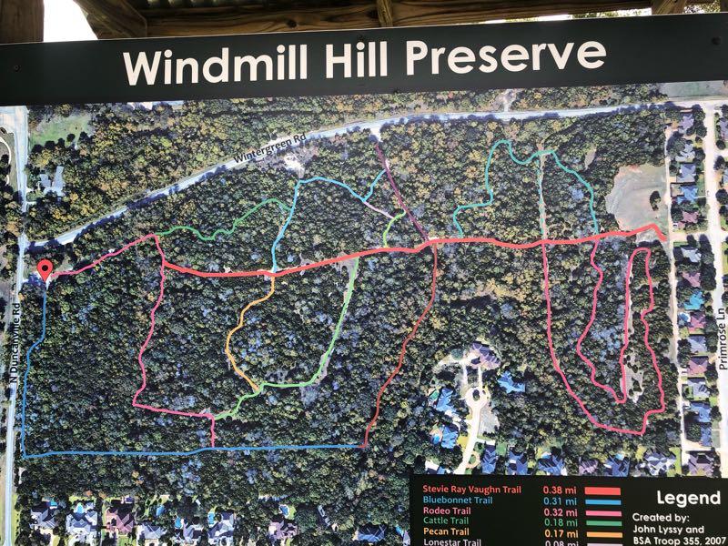 A detailed map of Windmill Hill Preserve showing various walking trails, including Stevie Ray Vaughn Trail, Bluebonnet Trail, Rodeo Trail, Cattle Trail, Pecan Trail, and Lenstar Trail. The map features color-coded paths in red, blue, green, and orange, along with a legend indicating the lengths of each trail. The surrounding area includes roads and residential homes. Windmill Hill Preserve mountain bike trail.