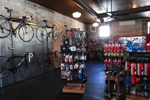 A bright bicycle shop interior featuring various bicycles mounted on a wall, with a display of bike accessories and tools on shelves. The space has exposed brick and metal elements, creating an industrial feel, while additional bike gear is visible in the background.