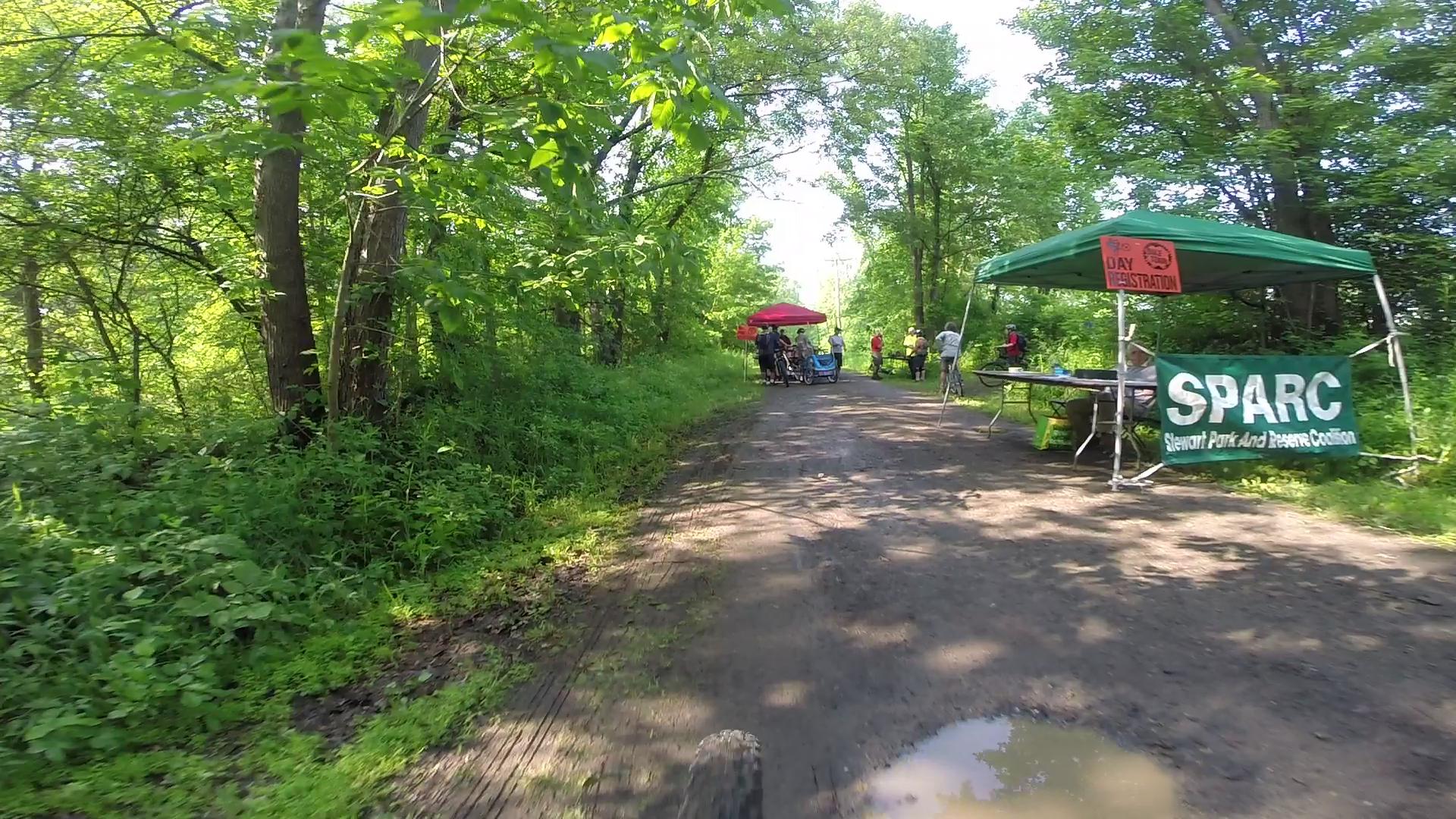 A dirt path surrounded by lush green trees, with a canopy set up for a registration area. Several people are gathered near the tent, which has a sign indicating "Day Registration" for SPARC (Stewart Park and Reserve Coalition). A muddy puddle is visible on the path, suggesting recent rain. Stewart State Forest mountain bike trail.