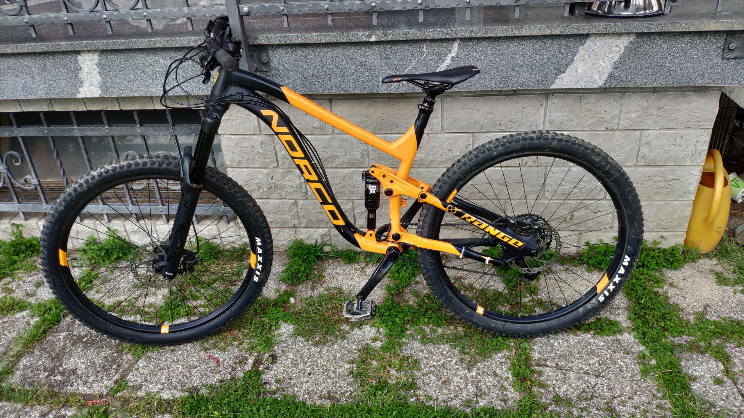 Norco Norco Range A7.3: A black and orange Norco mountain bike is parked on a gravel surface with green grass surrounding it. The bike features thick tires with "Maxxis" branding and a sturdy frame design. In the background, there is a stone wall and a decorative metal gate.