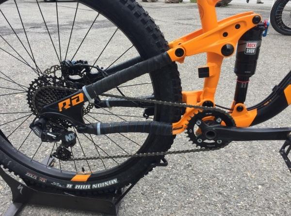 Norco Norco Range A7.3: Close-up of a mountain bike's rear drivetrain, showcasing the orange frame, chain, and gear components, with visible tire tread and suspension system. The image highlights the bike's intricate design and rugged build, suitable for off-road cycling.