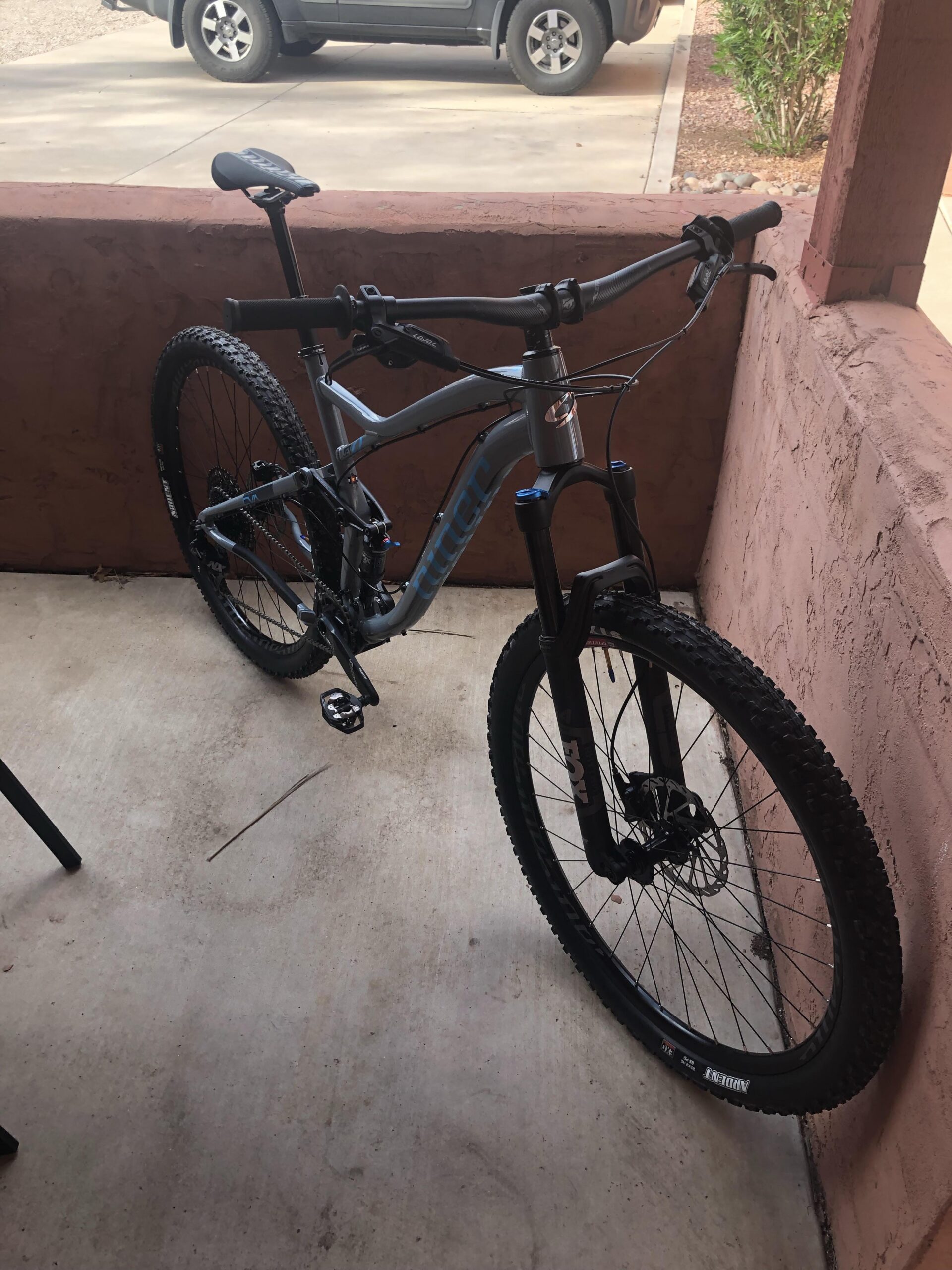 Niner Jet 9: A gray mountain bike is parked on a concrete surface next to a textured reddish wall, with a partially visible SUV in the background. The bike features thick tires and a front suspension fork.