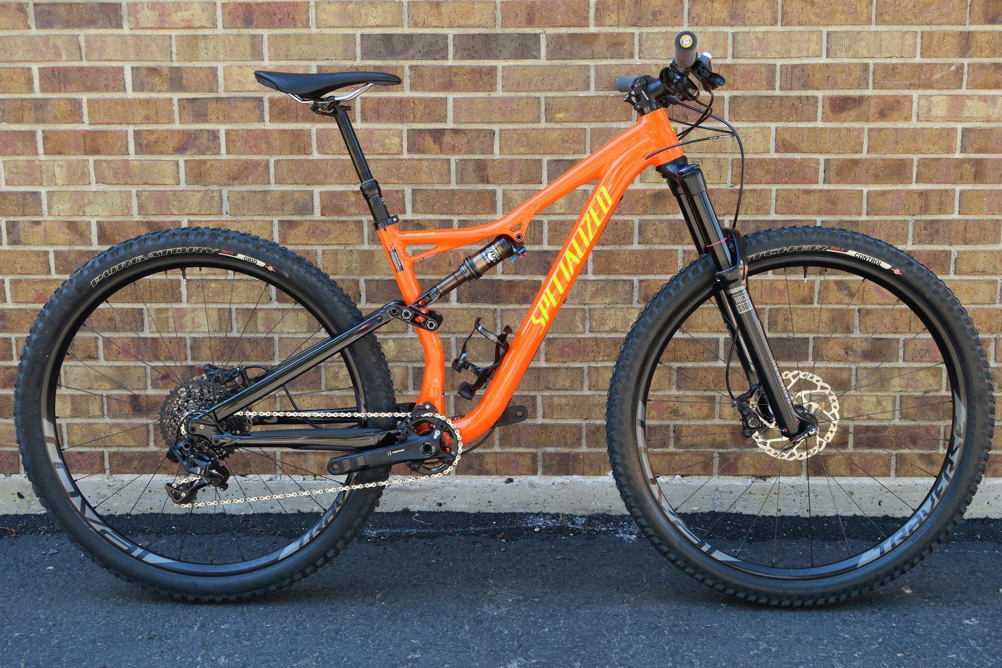 Specialized Stumpjumper FSR Elite 650b: A bright orange mountain bike with black components, featuring a lightweight frame, wide tires, and a suspension fork. The bike is positioned against a textured brick wall, highlighting its design and features.