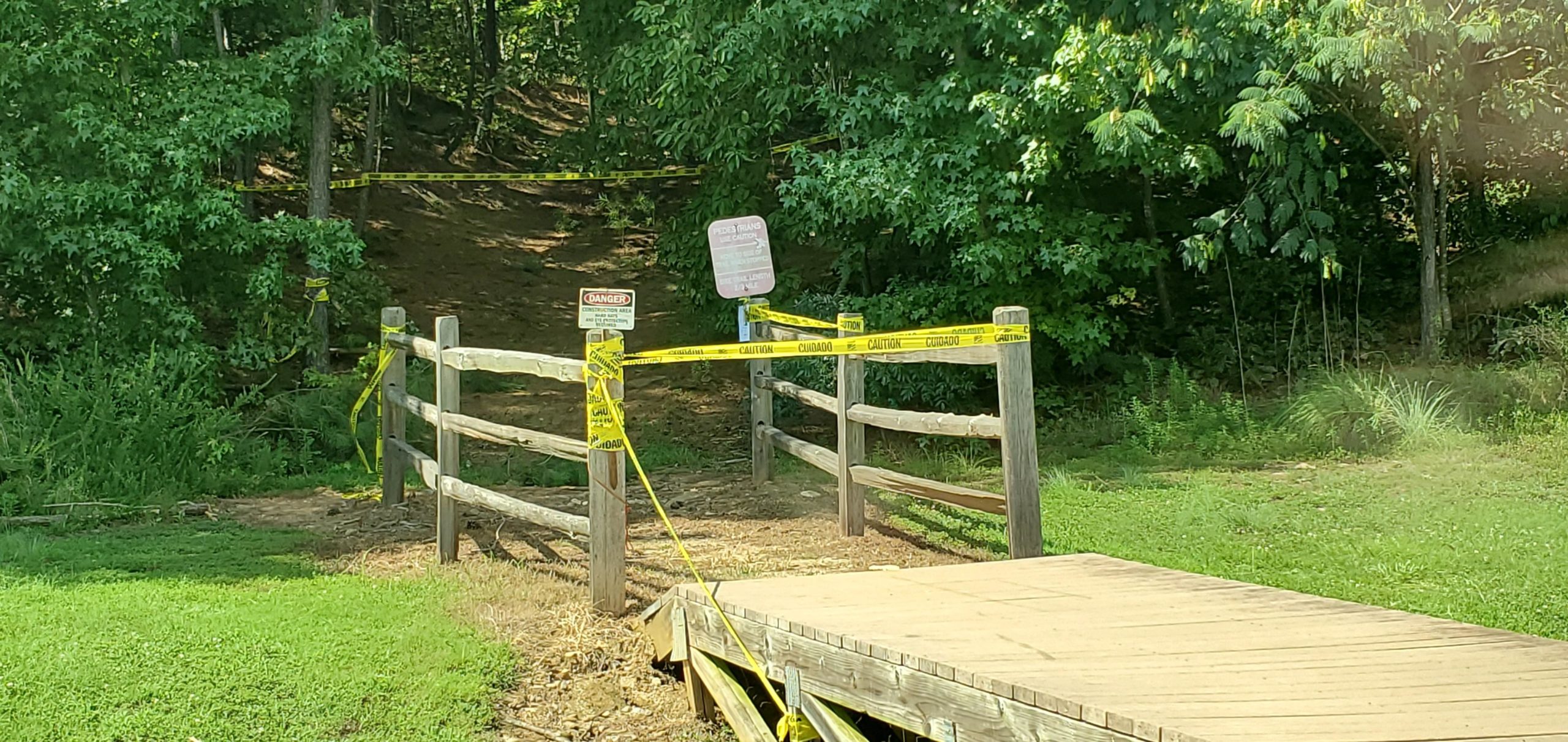 A wooden pathway leads to a fenced-off trail entrance marked with caution tape and signs indicating a dangerous area. Lush greenery surrounds the scene, with trees and vegetation visible on either side. North Cooper Lake Park Trail mountain bike trail.