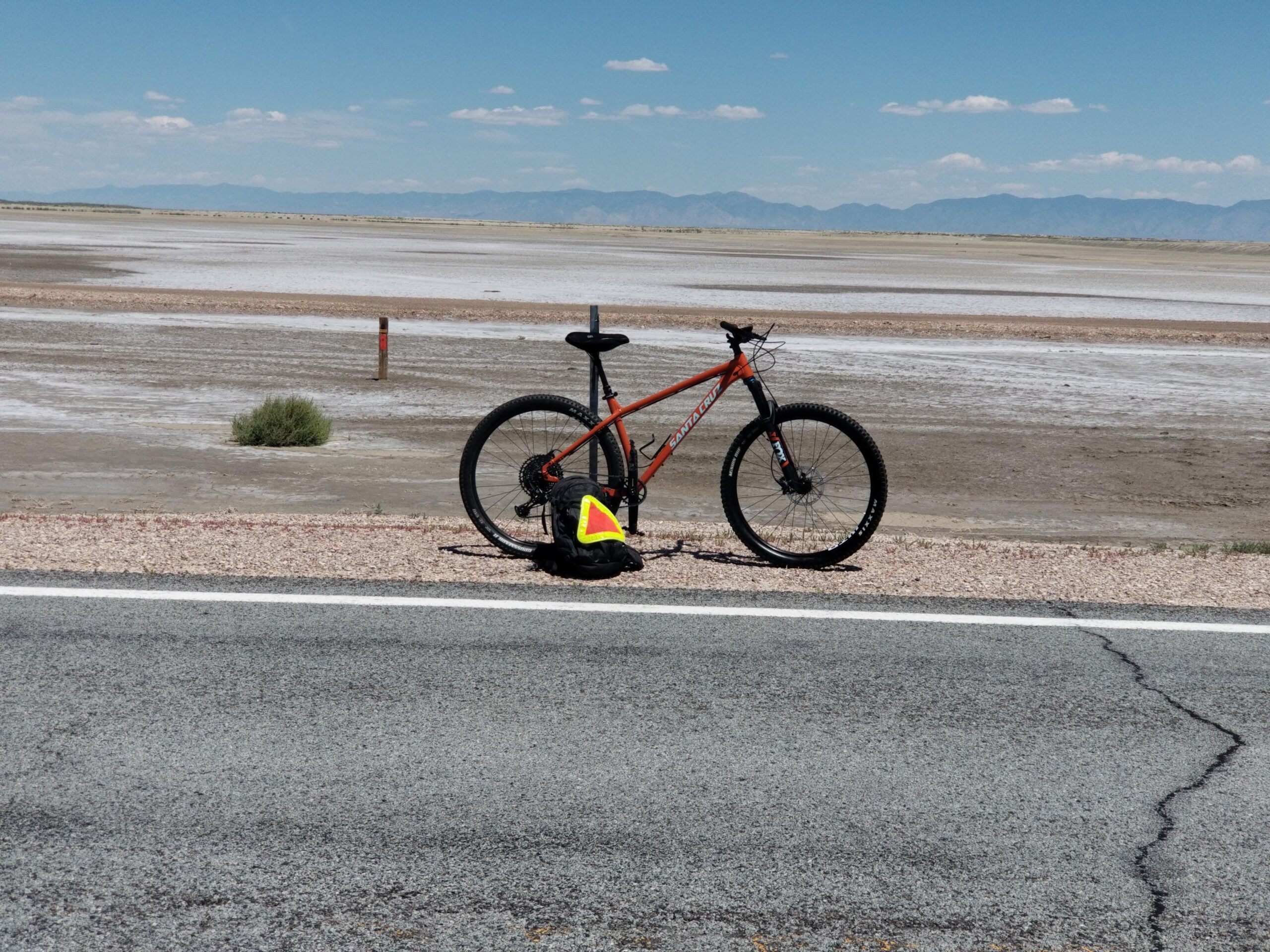 Santa Cruz Chameleon: A mountain bike with an orange frame is positioned near a dirt road, with a backpack beside it. In the background, a flat landscape stretches out, likely a dried salt flat, under a clear blue sky with scattered clouds and distant mountains on the horizon.