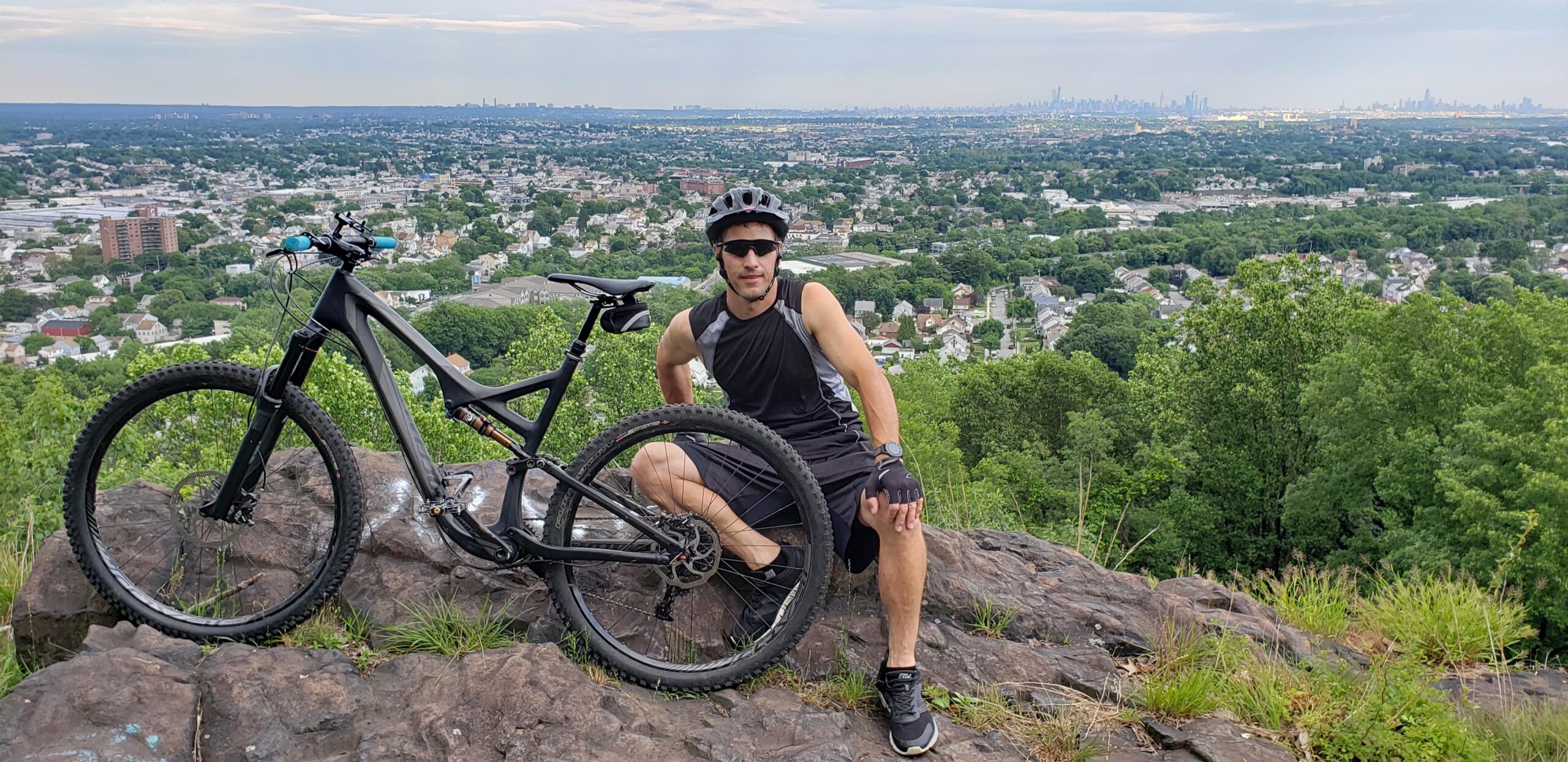 Specialized Stumpjumper FSR Comp EVO 29: A person wearing cycling gear kneels beside a mountain bike on a rocky outcrop. The background features a panoramic view of a green landscape and a city skyline in the distance under a cloudy sky.