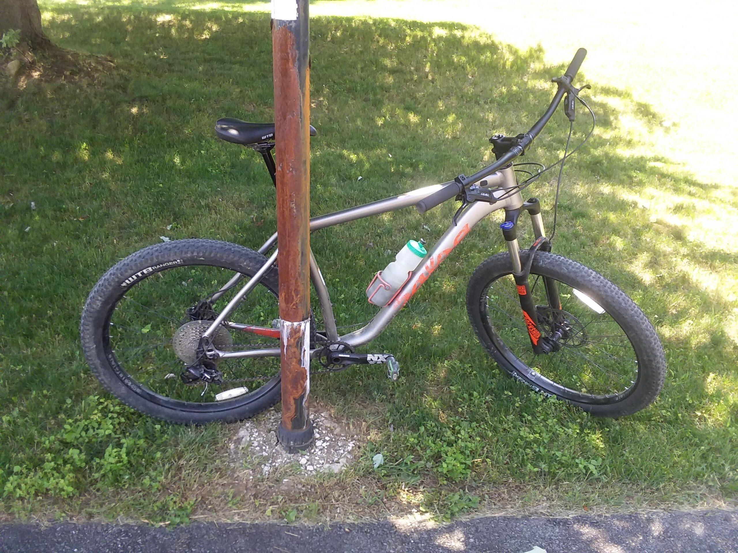 Salsa Timberjack: A mountain bike parked next to a weathered metal pole, set against a grassy background. The bike features thick tires, a water bottle attached to the frame, and a front suspension fork. Sunlight casts a soft glow, creating a relaxed outdoor atmosphere.