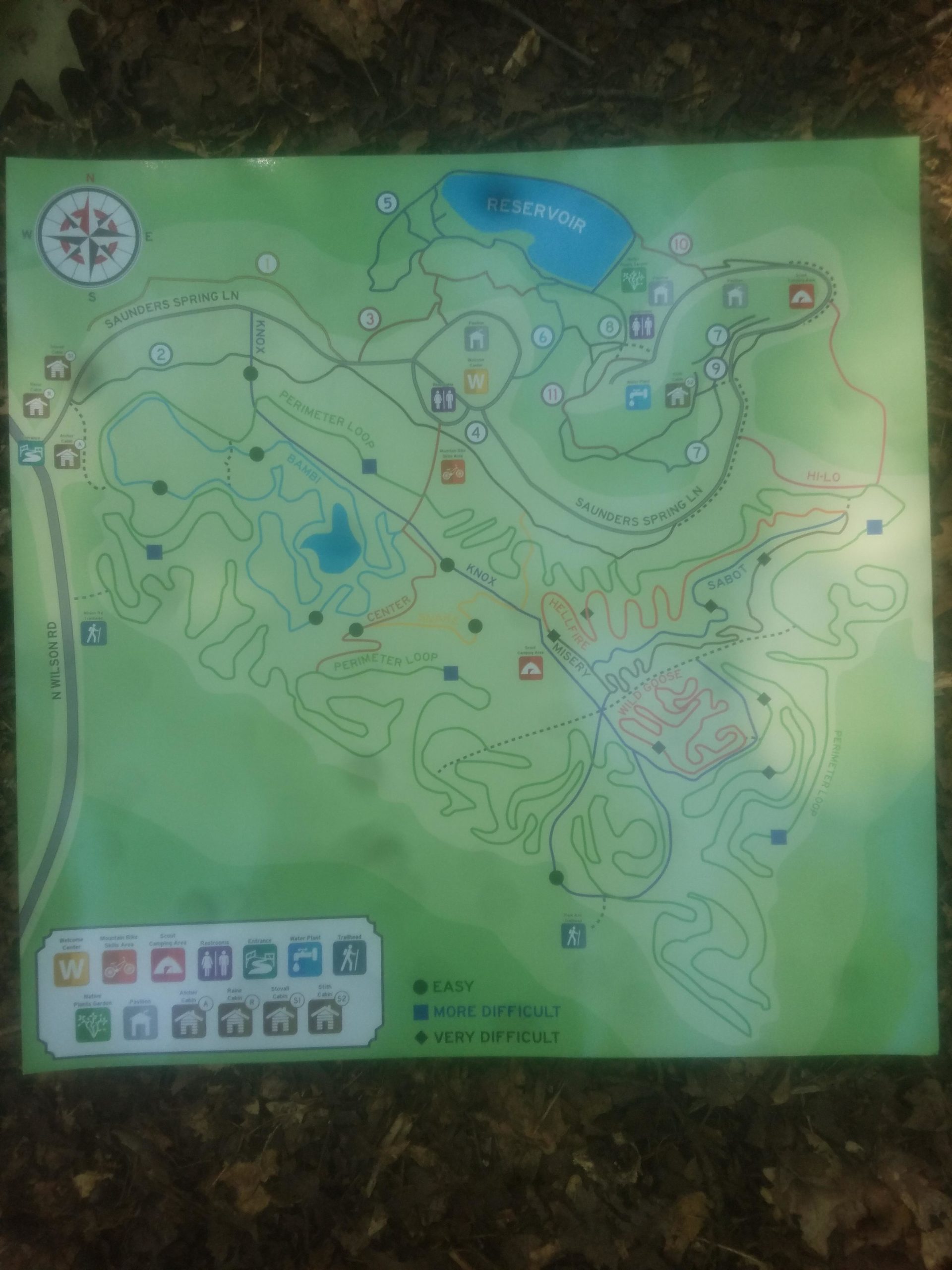 Map of a recreational area showing trails and difficulty levels, with a reservoir labeled at the top, a compass rose, and symbols for amenities. The trails are marked with color-coded difficulty levels: green for easy, blue for more difficult, and red for very difficult. Saunder Springs mountain bike trail.
