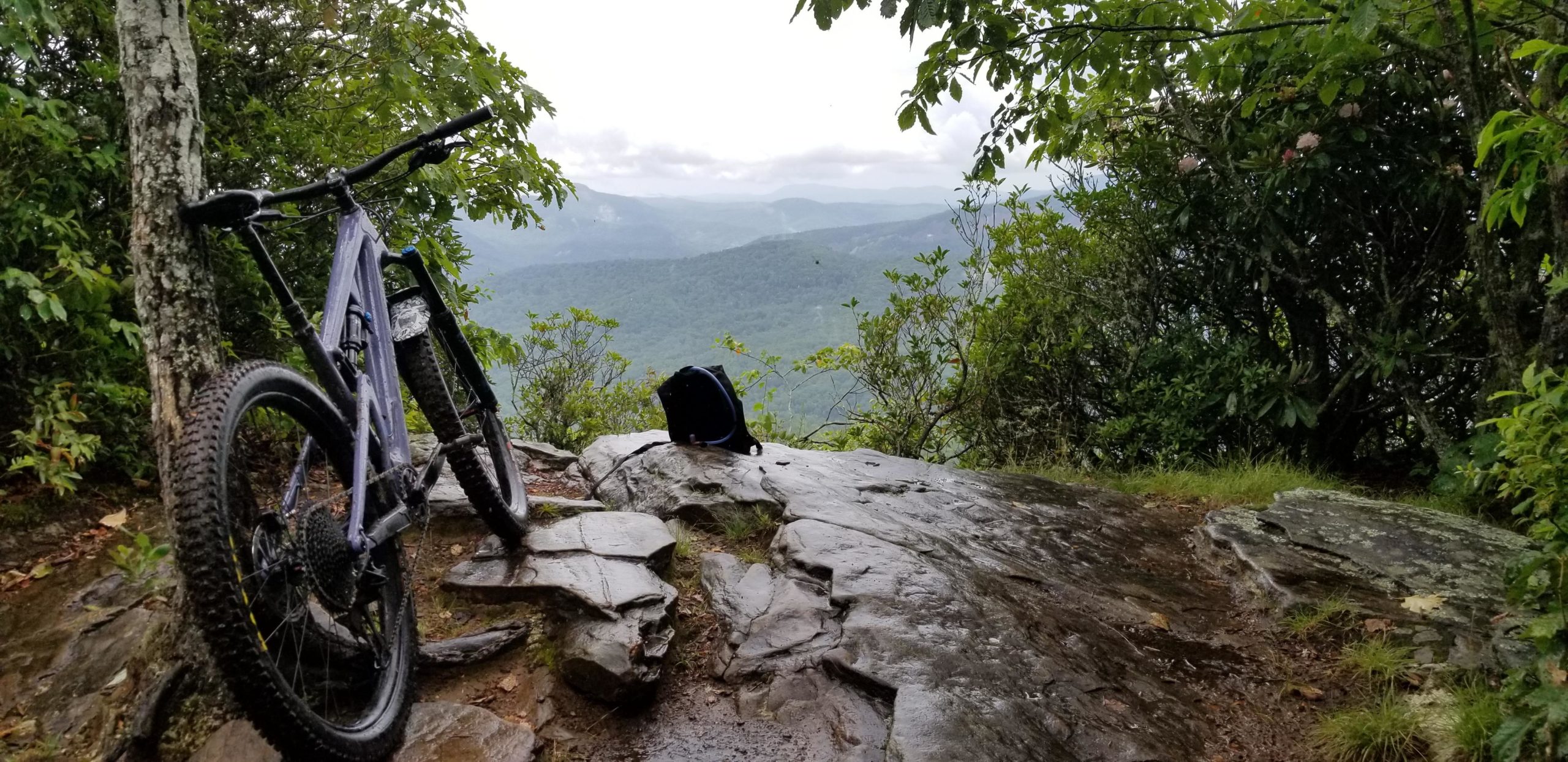 A mountain bike rests against a tree on a rocky outcrop, overlooking a lush green valley. The scene is slightly overcast, with distant mountains visible in the background. A small black bag sits on the rocks nearby, surrounded by foliage. Black Mountain mountain bike trail.