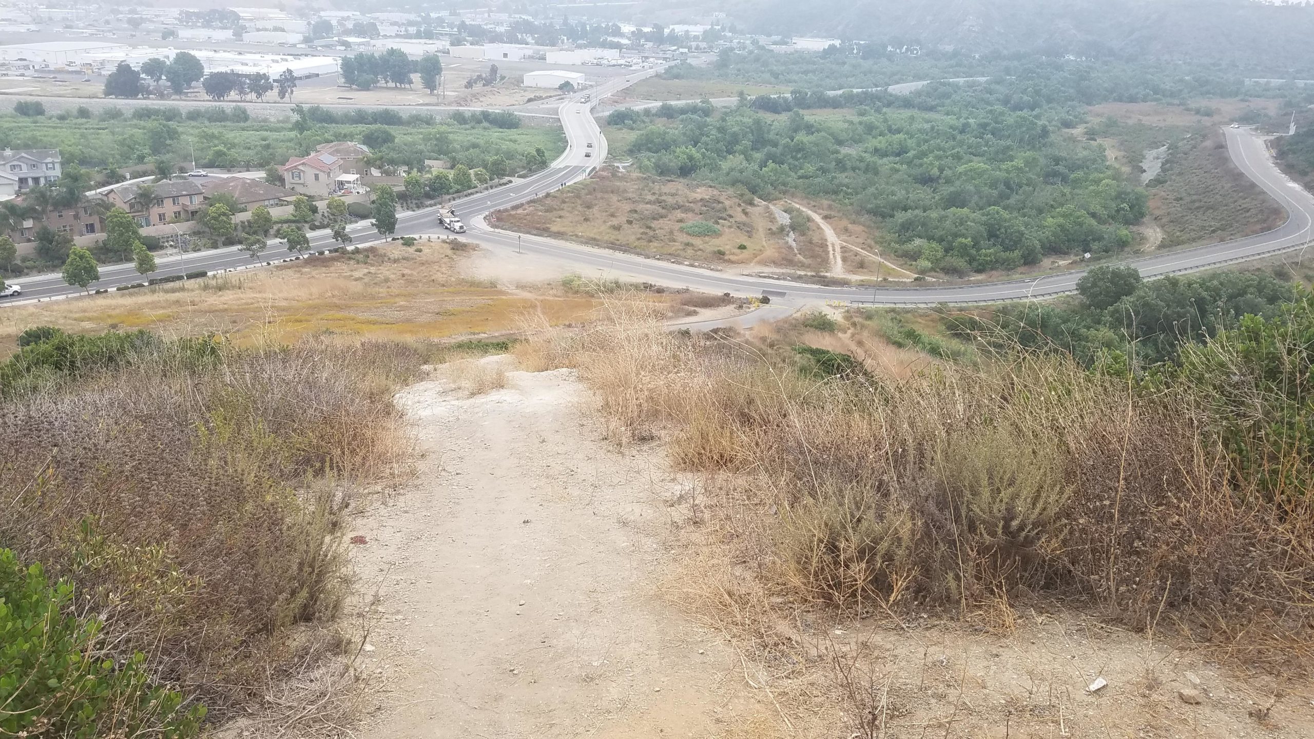 A view from a hillside overlooking a winding road with vehicles, residential homes, and a mix of greenery and dry vegetation in the foreground. The landscape features both developed areas and natural surroundings, with a distant view of hills in the background. Tuley Canyon Switchbacks mountain bike trail.