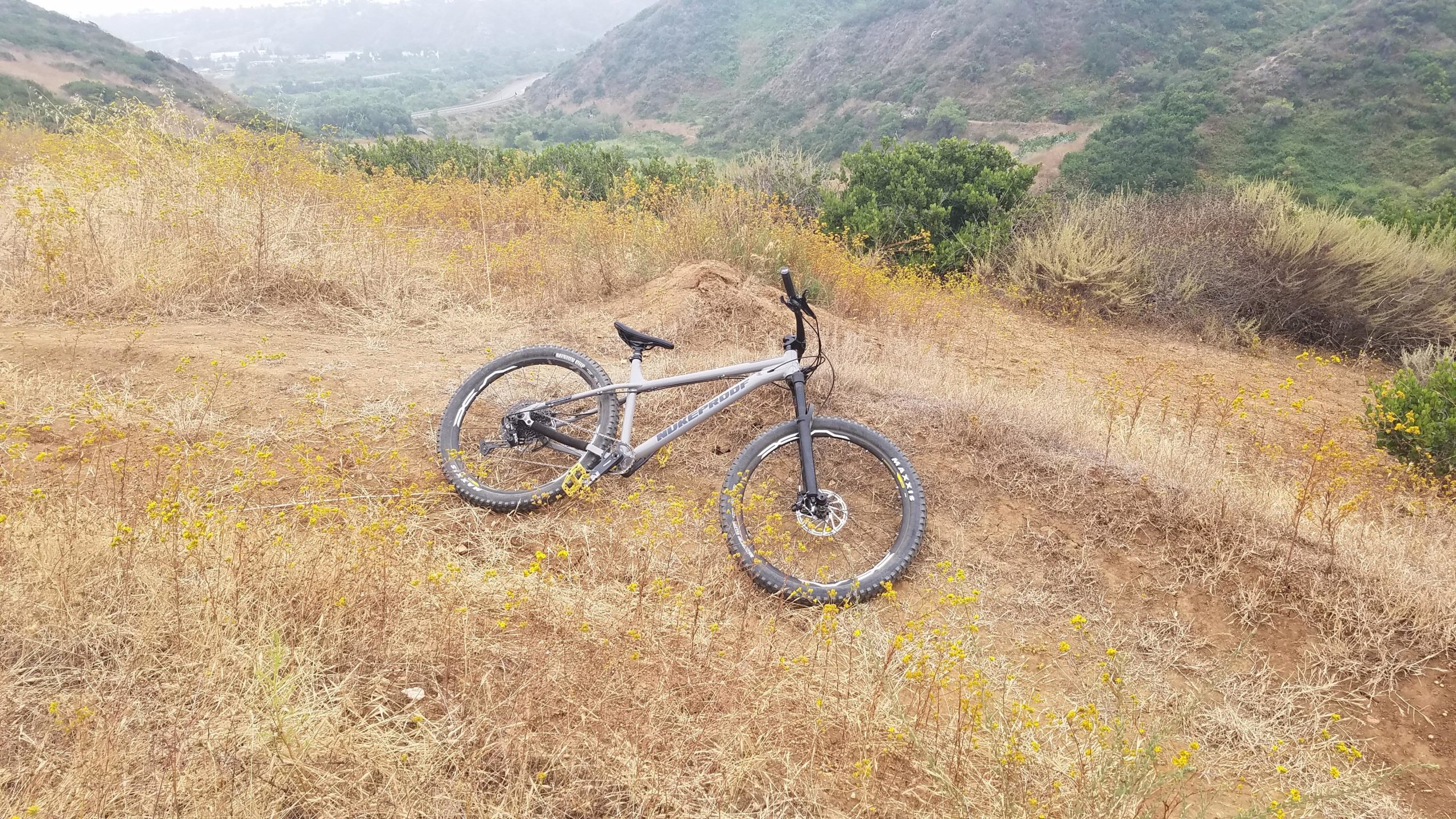 A silver mountain bike lies on its side in a dry, grassy area, surrounded by yellow wildflowers. In the background, rolling hills and a distant road can be seen, indicating a mountainous landscape. The atmosphere appears slightly misty, creating a serene outdoor scene. Tuley Canyon Switchbacks mountain bike trail.