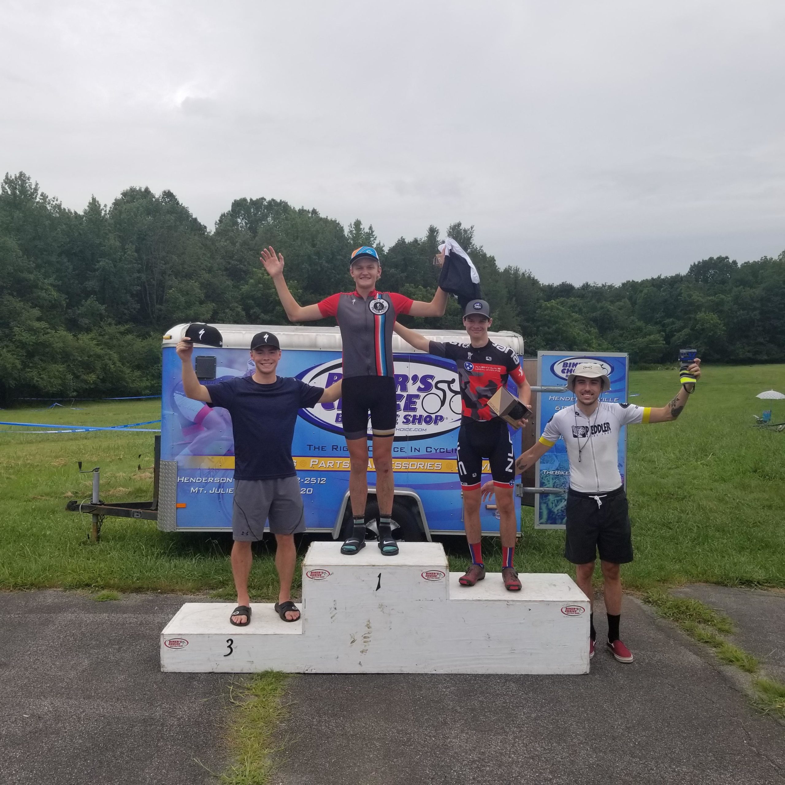 Four male cyclists celebrate on a podium after a race. The first-place winner, wearing a black and red cycling kit, raises his arms joyfully while holding a trophy. The second and third-place cyclists stand beside him, smiling and holding their awards, while the fourth cyclist gestures excitedly from the ground. In the background, there is a trailer with a bike shop logo, and a grassy field is visible under a cloudy sky. Lock 4 mountain bike trail.