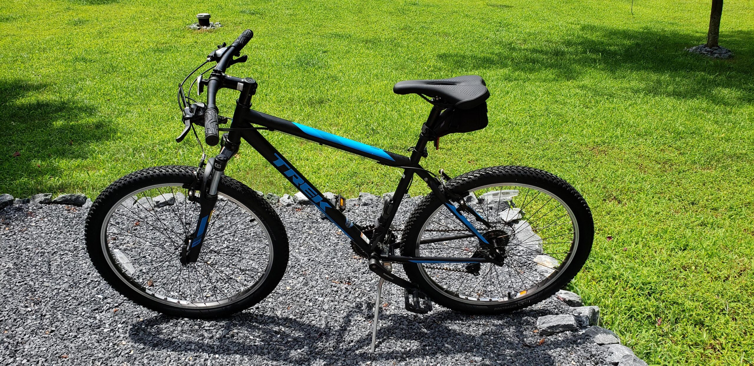 Trek 820: A black and blue Trek mountain bike parked on gravel with green grass in the background. The bike features a standard design for mountain biking, with thick tires, a front suspension fork, and a black seat bag.
