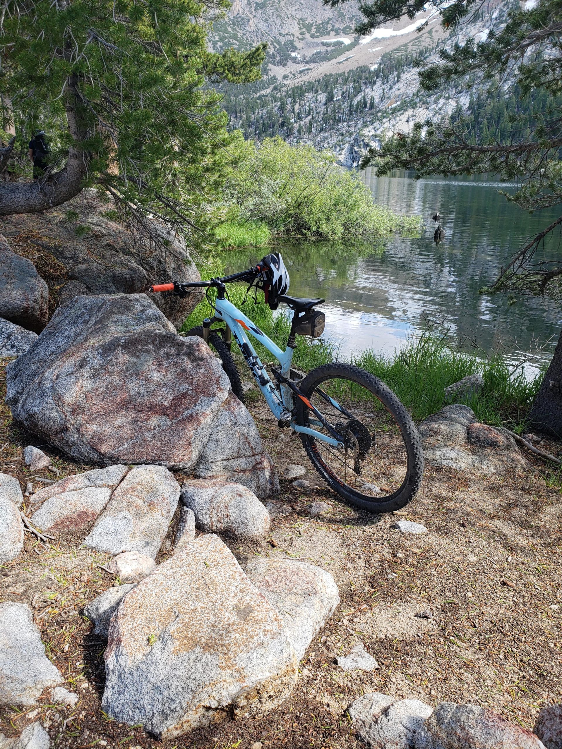 A mountain bike resting against large rocks near a serene lake, surrounded by lush greenery and pine trees, with a mountainous landscape in the background under clear skies. Star Lake Trail mountain bike trail.