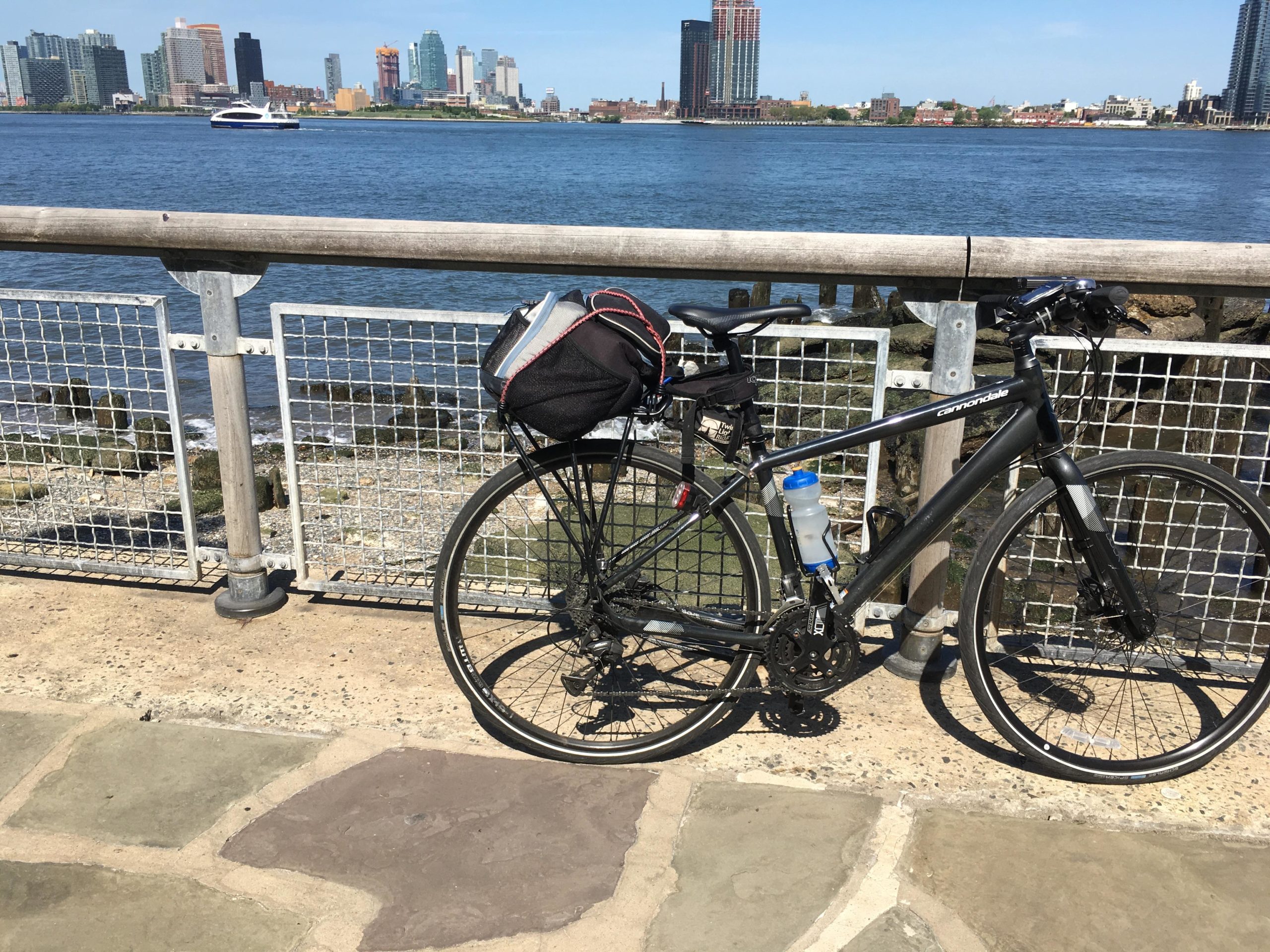 A parked black Cannondale bicycle with a water bottle attached, situated beside a waterfront path with a railing. The background features a view of a city skyline across the water on a sunny day. East Side Green way 34th st to the Staten Island Ferry mountain bike trail.