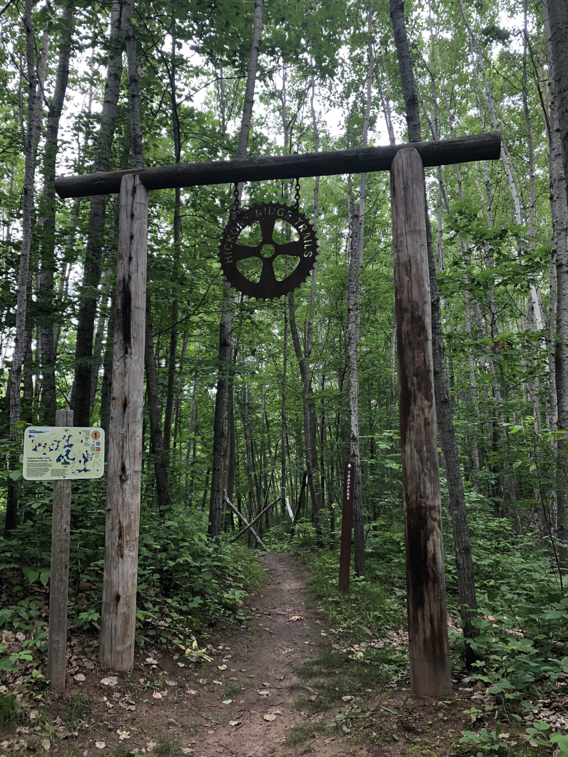 A wooden entrance arch with a gear-shaped sign reading "Hickory Ridge Trails" is seen at the beginning of a forested trail. Flanking the entrance are two posts, one with a trail map and the other indicating trail directions. The path leads into a lush green forest with tall trees and thick foliage. Hickory Ridge mountain bike trail.