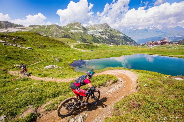 A scenic view of mountain bikers navigating a winding dirt trail through lush green hills, with a clear blue lake and distant mountains in the background under a partly cloudy sky.
