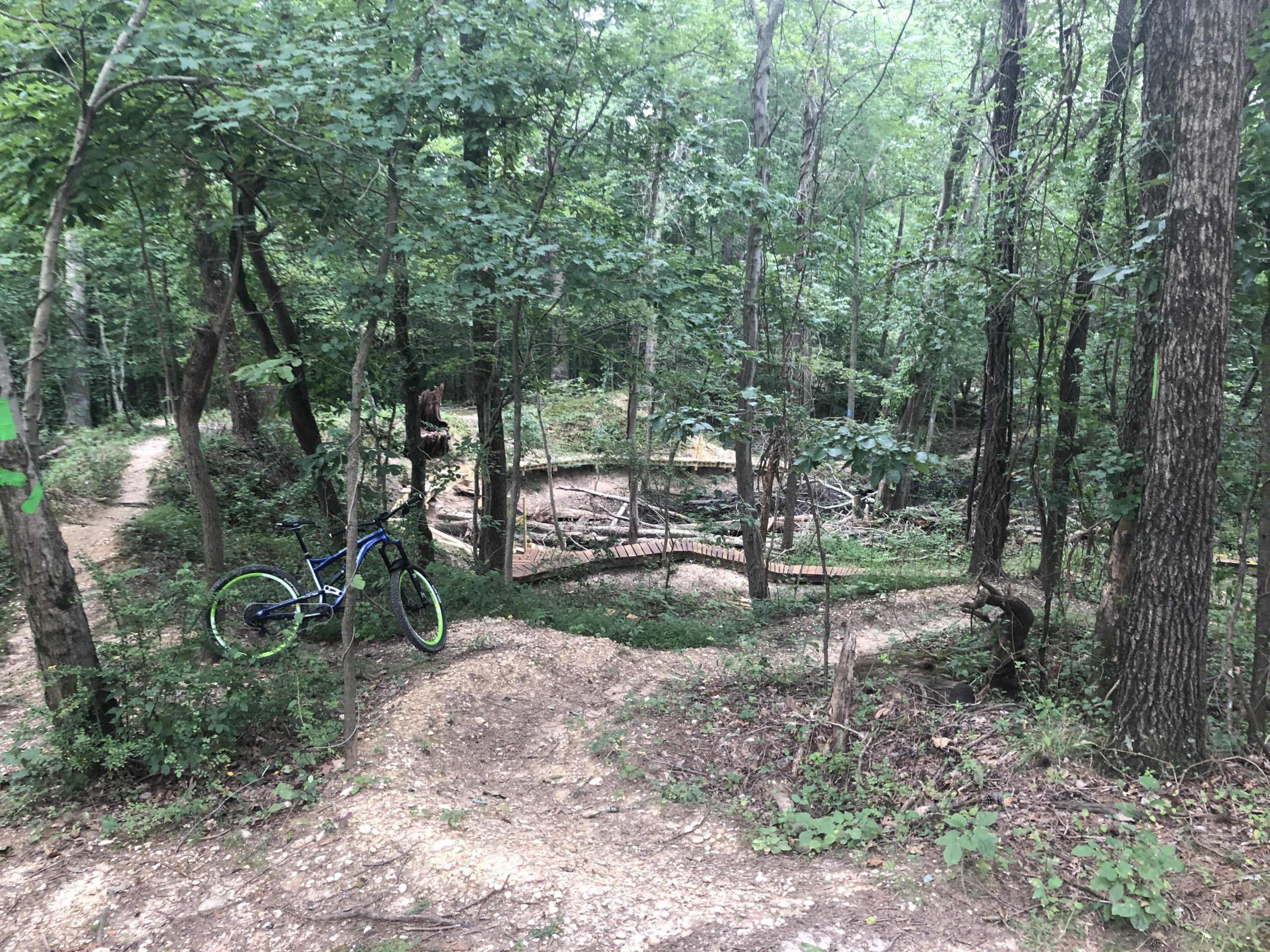 A mountain bike resting on a gravel path surrounded by lush green trees in a wooded area, with winding trails visible in the background. Fairland Recreational Park mountain bike trail.