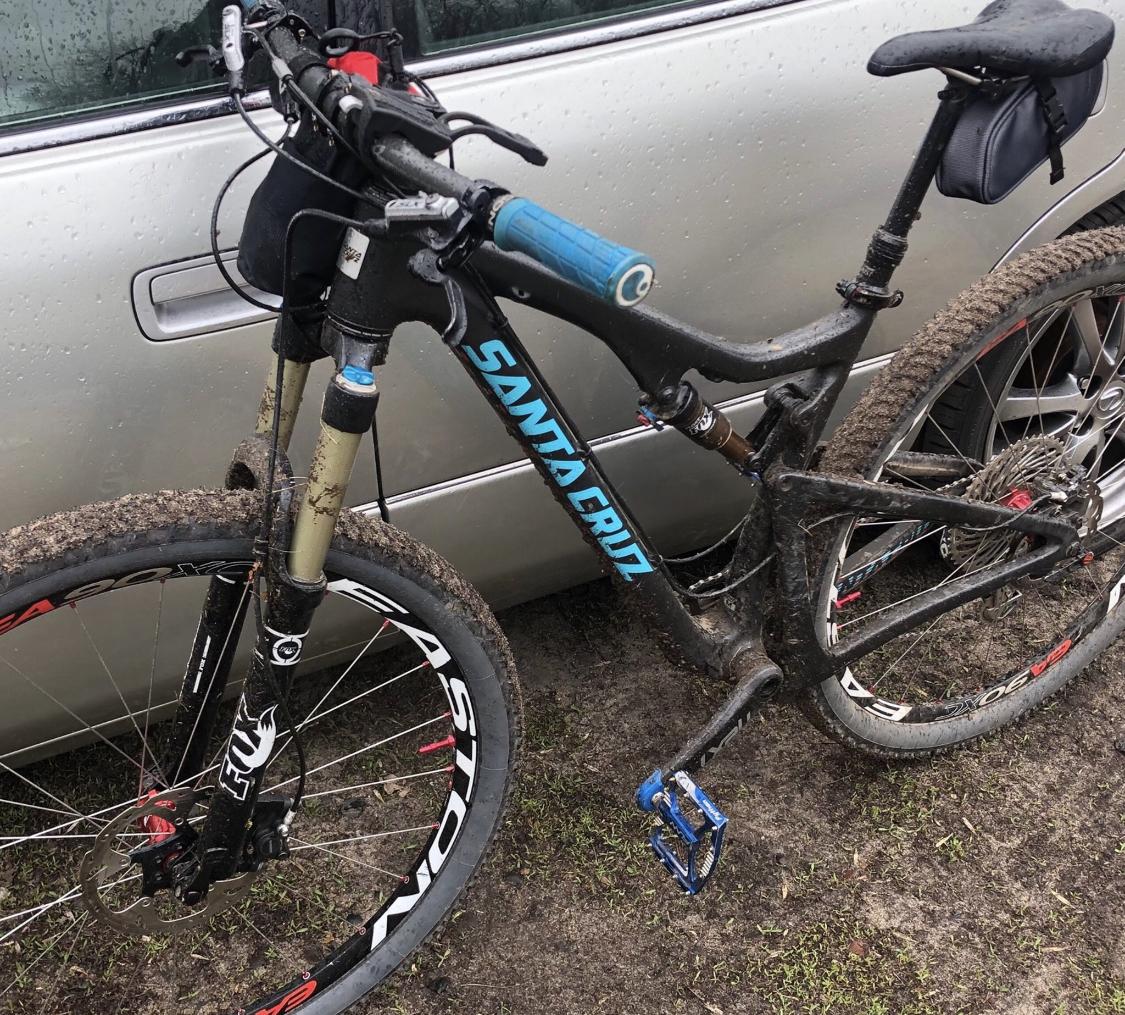 Santa Cruz Tallboy Carbon: A close-up view of a muddy Santa Cruz mountain bike leaning against a silver car. The bike features a black frame with blue accents, a suspension fork, and dirty tires. The handlebars have blue grips, and there is a small black bag attached to the frame. The background shows wet ground with grass.