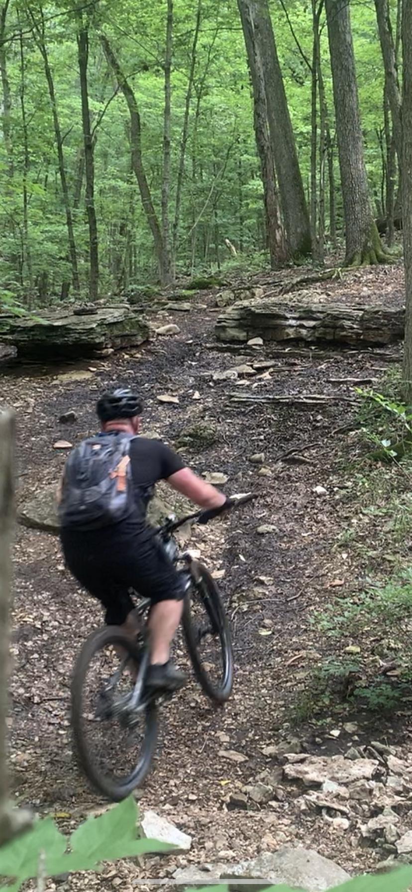A person biking on a rocky, forested trail surrounded by lush green trees. The biker is wearing a helmet and a backpack, and is navigating a path with uneven surfaces and large rocks. Chubb Trail mountain bike trail.