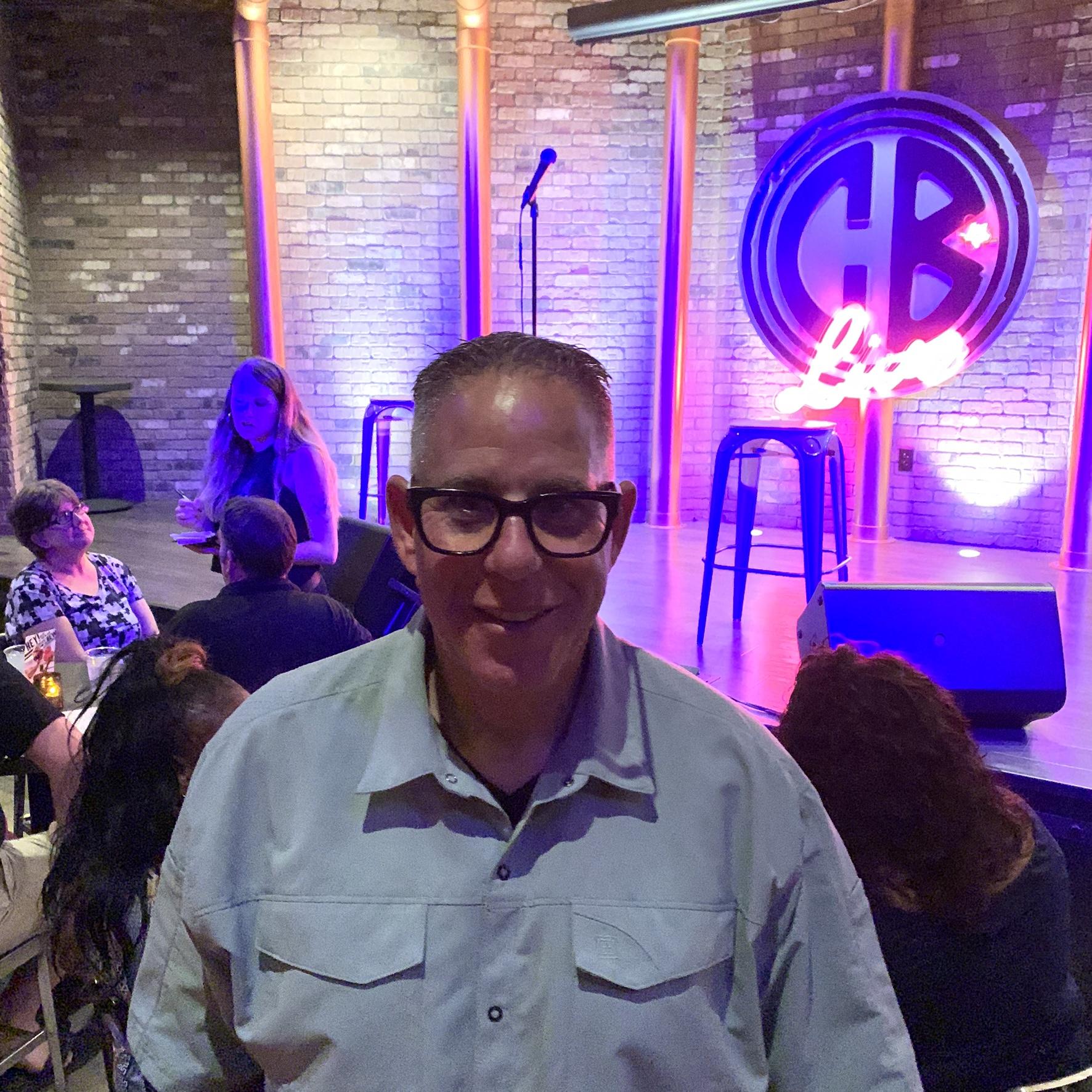 Pivot Mach 5.5: A smiling man with glasses is in the foreground of a lively comedy club scene, with a colorful neon sign reading "CB Live" in the background. There are people seated at tables, and a performer is on stage. The setting features a brick wall and stage lighting, creating an atmospheric vibe for the event.