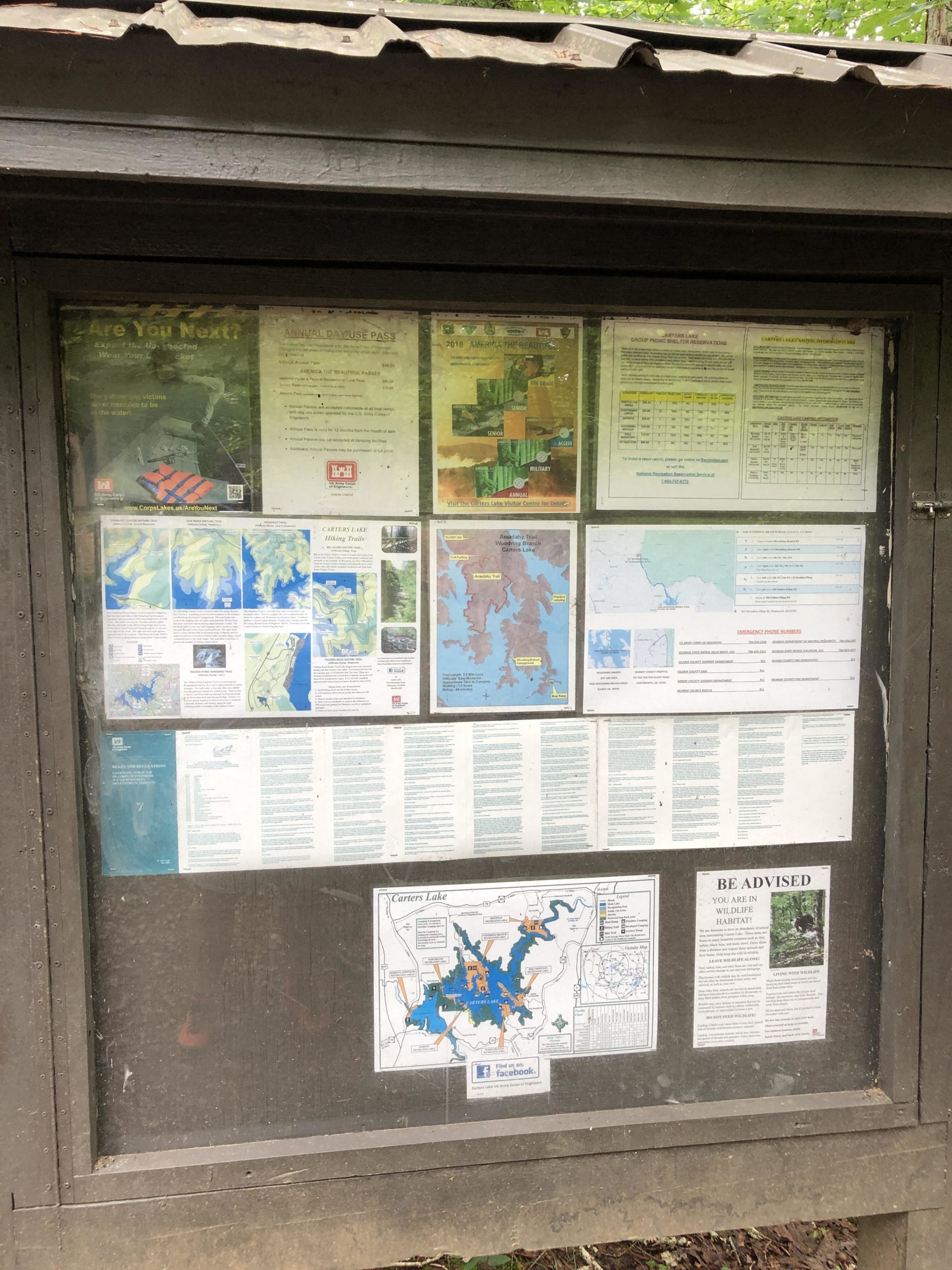 Information display board featuring various maps and notices related to recreation and safety at a park, including trail maps, wildlife habitat advisories, and pass information. The board is enclosed in a wooden structure with a corrugated roof and is located in a forested area. Woodring Branch mountain bike trail.