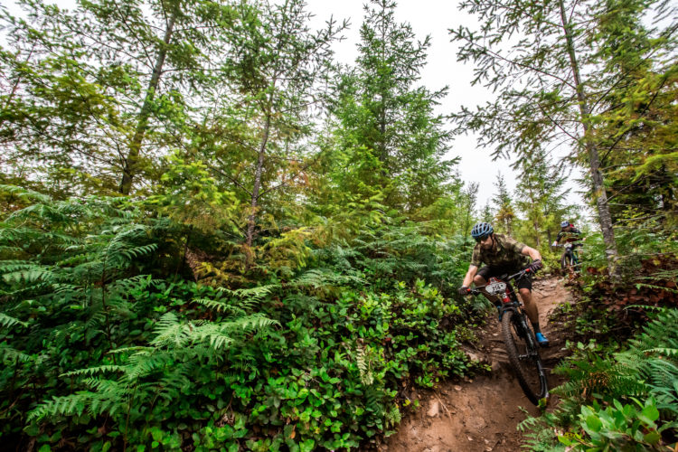 A mountain biker navigating a dirt trail surrounded by lush greenery and tall trees, with another cyclist visible in the background. The scene captures the thrill of outdoor biking in a forested area.
