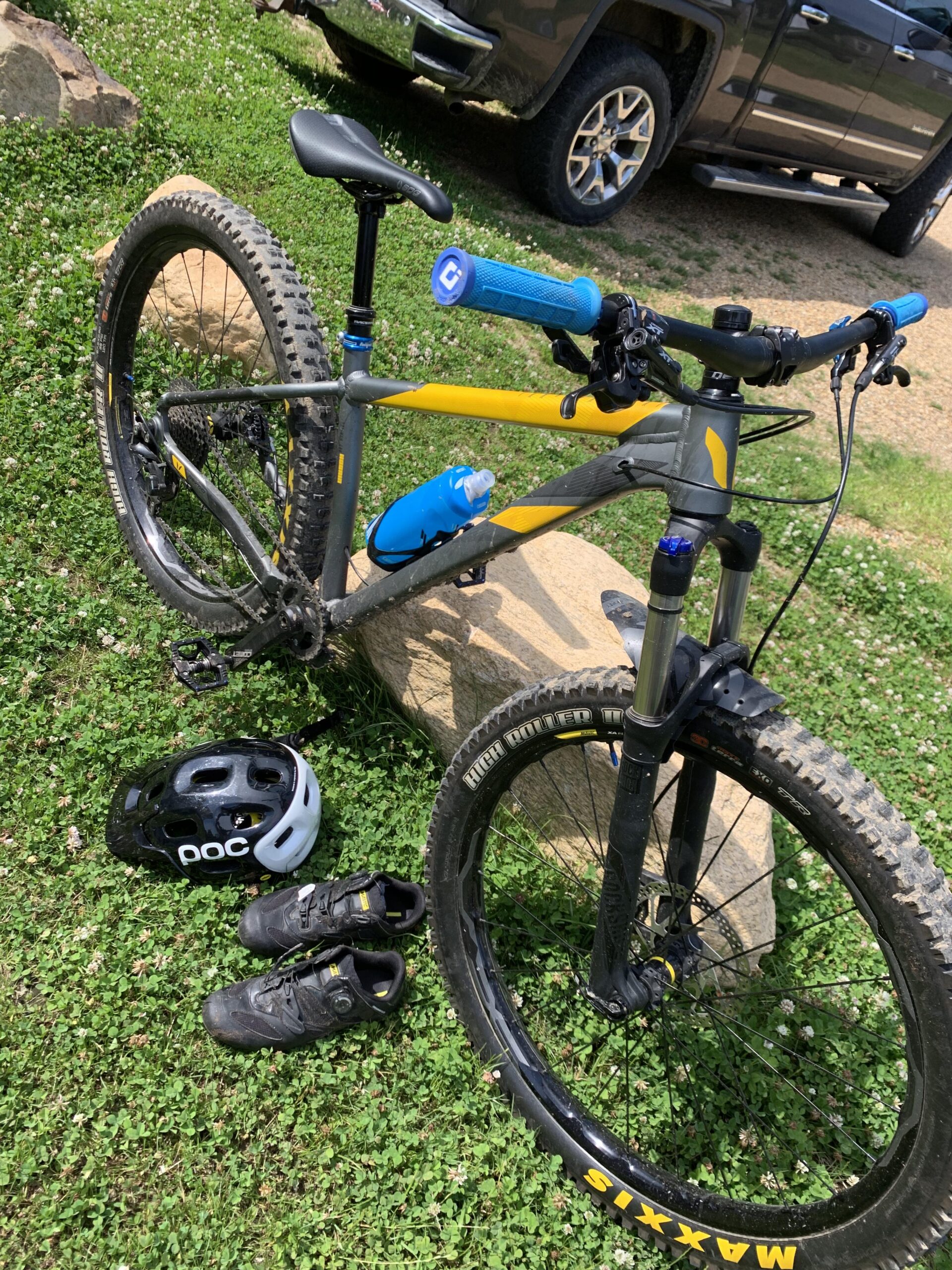 Devinci Devinci Kobain: A mountain bike with a gray and yellow frame is parked on a grassy area next to a large rock. There is a blue water bottle attached to the frame, and blue handlebar grips. A black helmet and a pair of black cycling shoes are placed on the ground nearby. In the background, a pickup truck is partially visible.
