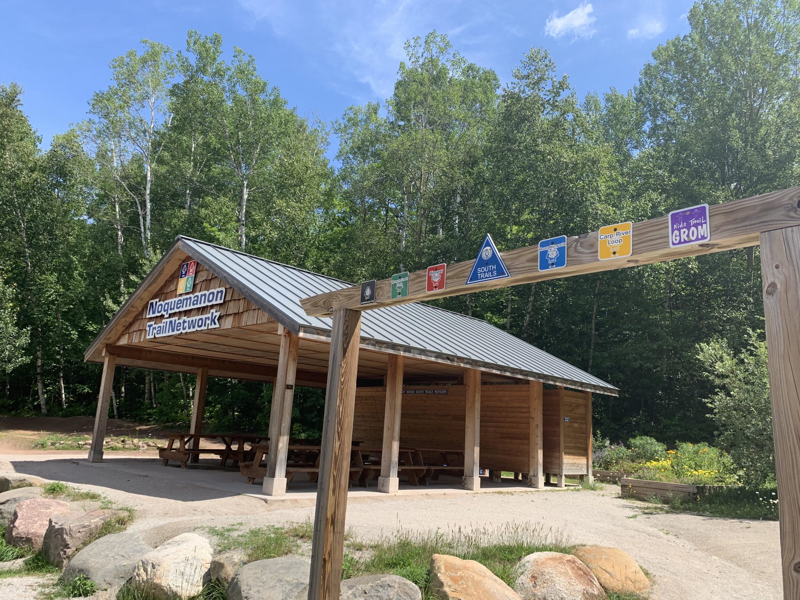 A wooden shelter with a metal roof marked "Noquemanon Trail Network" stands in a natural area surrounded by trees. A signpost with various trail markers, including symbols for different trail routes, is visible in the foreground. The scene is set under a clear blue sky, highlighting the lush greenery of the surrounding landscape. Noquemanon Trails Network: South Marquette Trails mountain bike trail.
