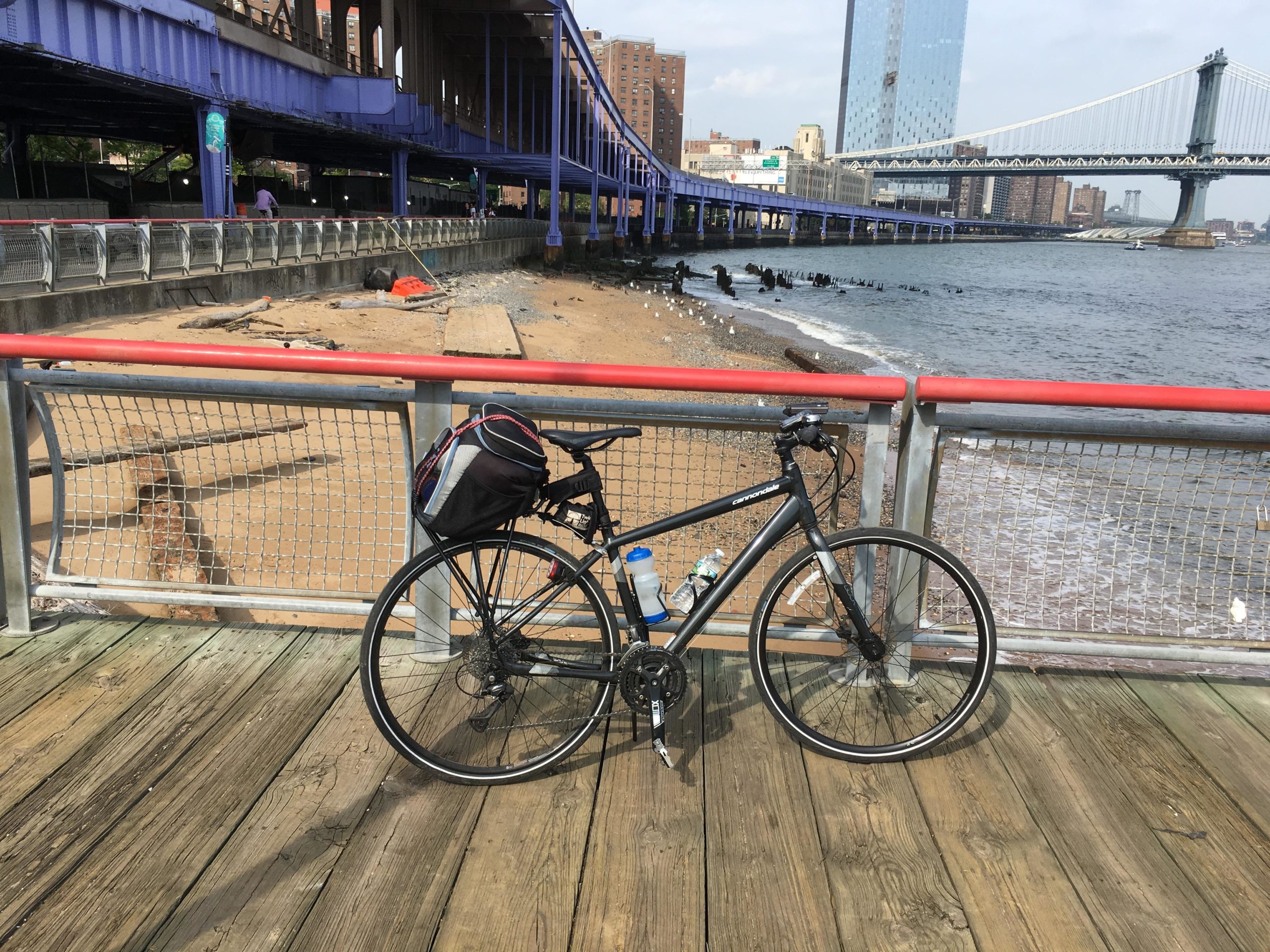 A black bicycle with a water bottle attached rests against a railing on a wooden platform by a riverside. In the background, a sandy area is visible, along with a bridge and city buildings. The sky is partly cloudy, and the water shows gentle waves. East Side Green way 34th st to the Staten Island Ferry mountain bike trail.