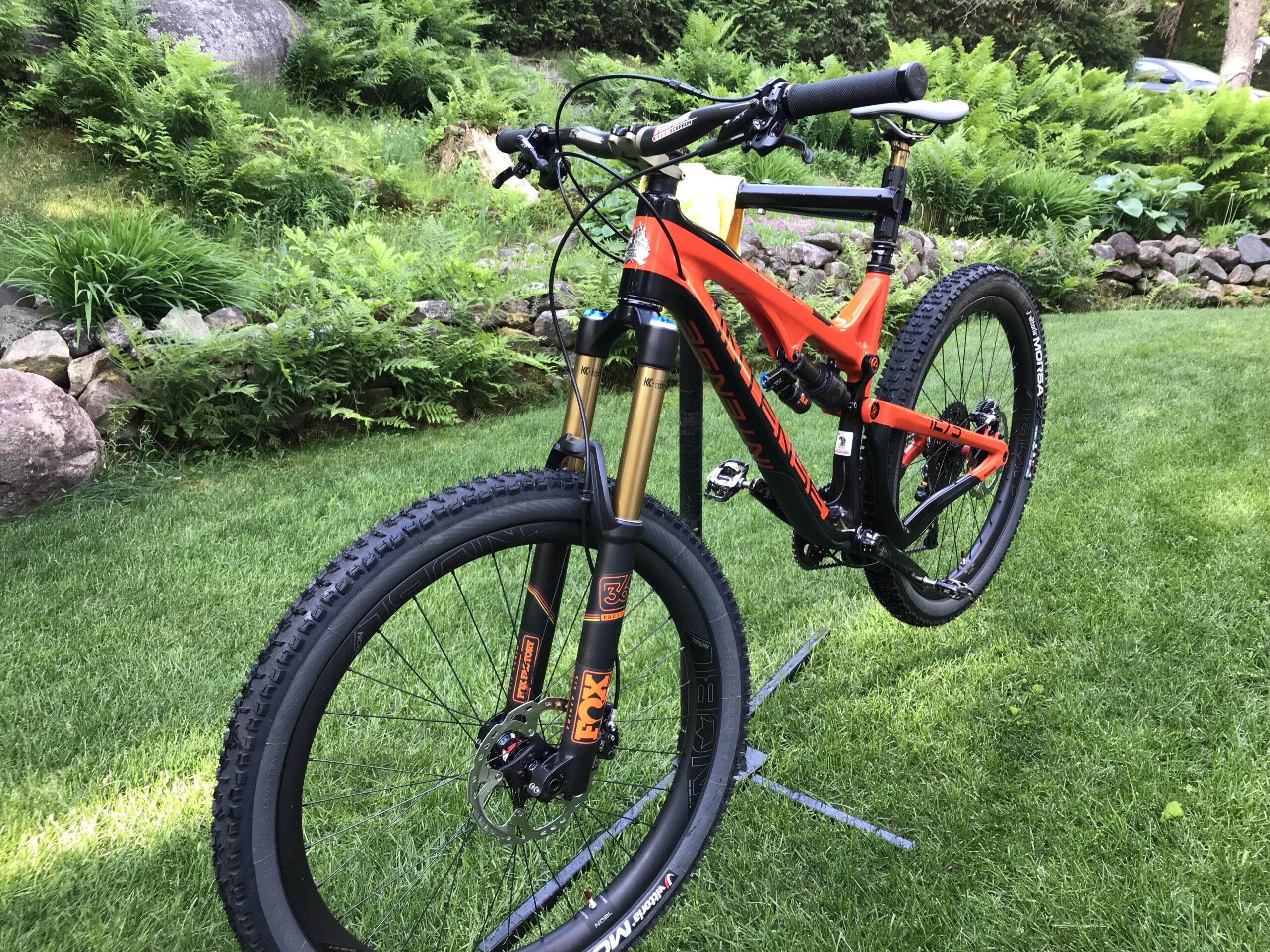 Intense Tracer 2: A close-up view of a mountain bike featuring a bright orange and black frame, positioned on a grassy area. The bike is equipped with high-performance components, including a suspension fork and rugged tires suitable for off-road riding. In the background, there is a lush green landscape with ferns and rocks.