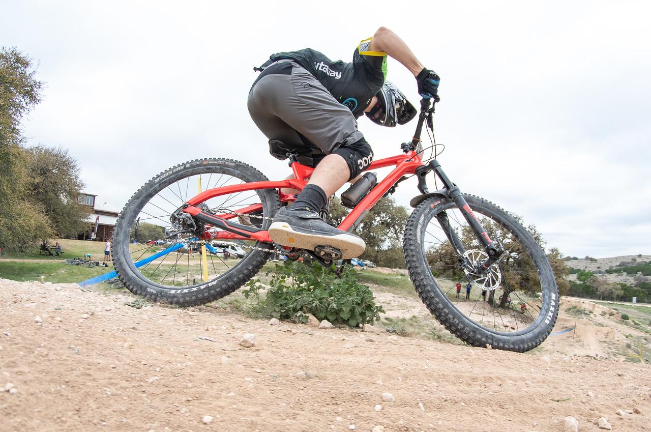 Devinci Troy Carbon RR: A mountain biker performing a jump on a red bike, with a dirt path and trees in the background. The rider is wearing a helmet, gloves, and protective gear, showcasing an active outdoor scene.