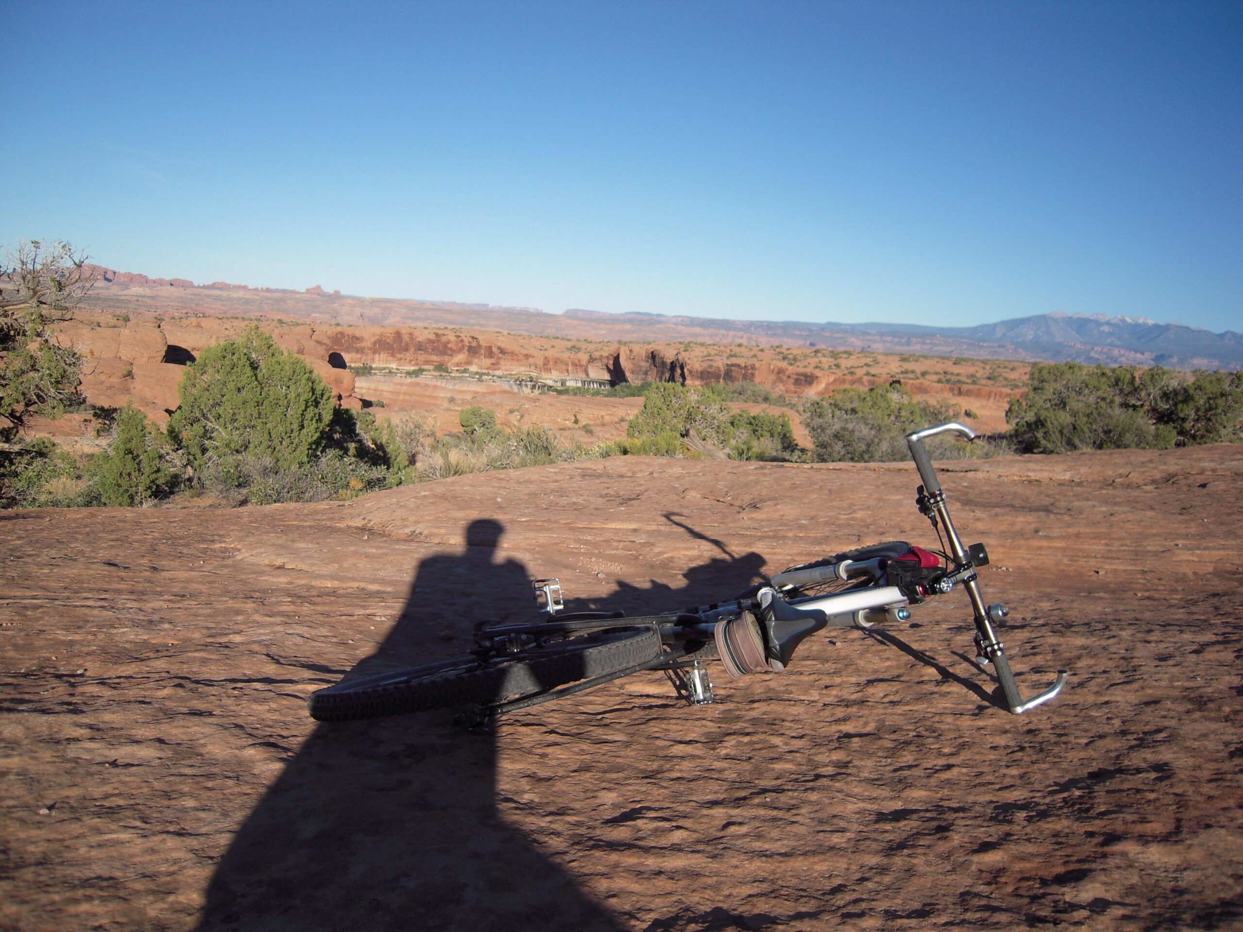 Specialized Epic Comp 29er: A mountain bike resting on a rocky landscape, with expansive views of distant cliffs and a clear blue sky. The shadow of the bike's rider is visible on the ground, adding depth to the scene.