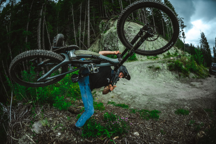 A mountain biker performing a maneuver with their bike, lifting it above their head while standing on a dirt path surrounded by greenery and trees. The scene depicts a dynamic outdoor setting with a cloudy sky in the background.