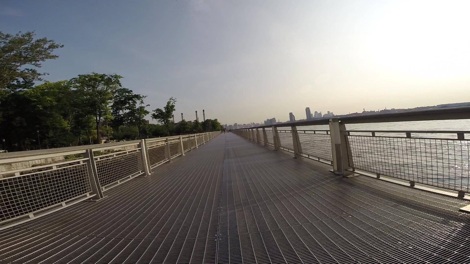 A sunlit pathway along a waterfront, featuring a metal grating surface and railing, with greenery on the left and a view of the city skyline in the background. East Side Green way 34th st to the Staten Island Ferry mountain bike trail.