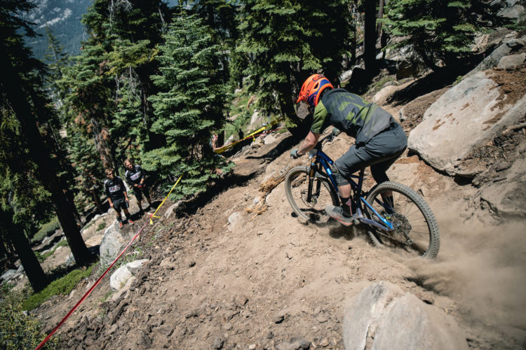 A mountain biker navigates a rocky trail in a forested area, kicking up dust as he rides down a slope. Two spectators watch from the side of the trail, surrounded by tall pine trees and rocky terrain. Yellow and red tape marks the boundaries of the course.