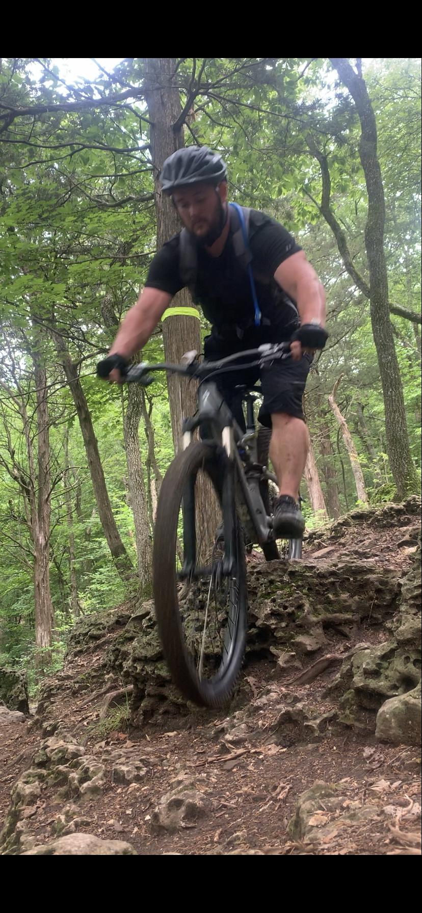 A person riding a mountain bike over rocky terrain in a forested trail, surrounded by trees and greenery. The rider is wearing a helmet and dark clothing, with a focus on navigating the uneven ground. Chubb Trail mountain bike trail.