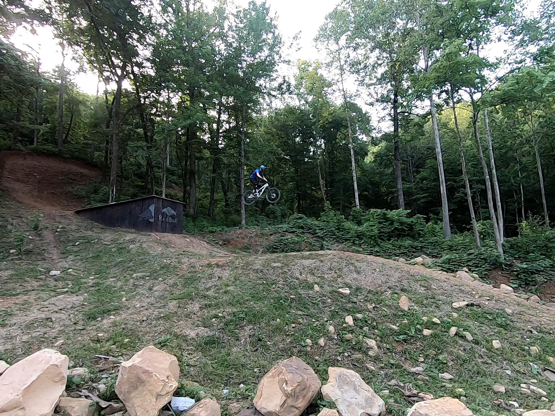 A person on a mountain bike performs a jump off a dirt ramp in a wooded area, with trees and greenery surrounding the scene. The ramp features a Red Bull logo, and rocks are visible in the foreground. Windrock Bike Park mountain bike trail.