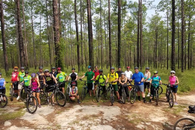 A diverse group of cyclists posing for a photo in a forested area, surrounded by tall pine trees. The group is wearing various colored biking jerseys and helmets, and each person is holding a mountain bike. The scene captures a sense of teamwork and outdoor adventure.
