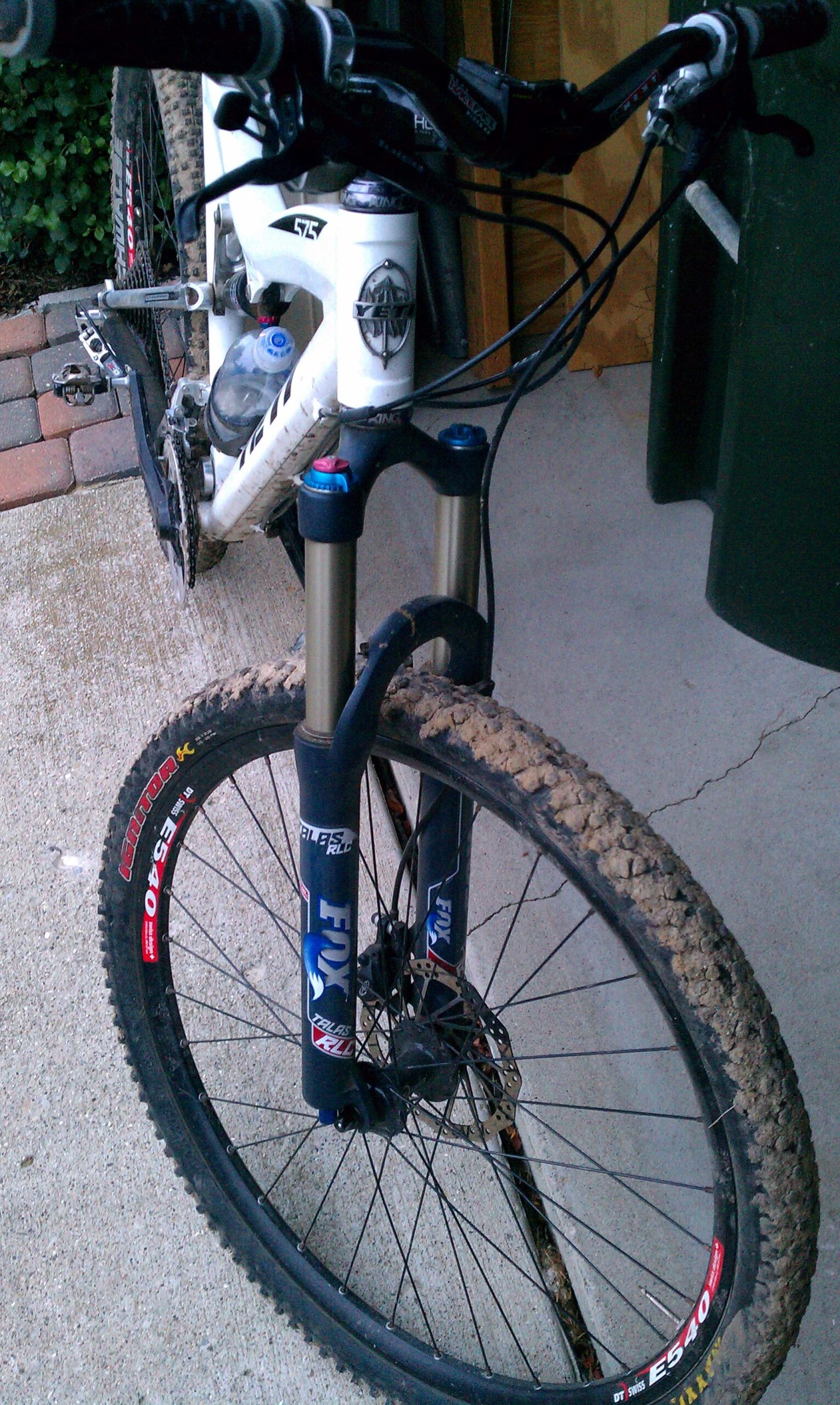 Yeti 575: Close-up view of a mountain bike's front fork and wheel, showcasing muddy tires and a water bottle mounted on the frame. The bike is parked on a concrete surface with a textured background, featuring a mix of gravel and grass.