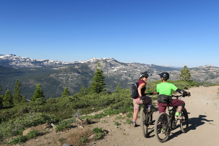 Two mountain bikers pause on a dirt trail, overlooking a scenic landscape with mountains and trees. One biker, wearing a bright green shirt and black shorts, is on the right, while the other, dressed in a dark top and colorful shorts, is on the left. The clear blue sky and snow-capped peaks in the background create a picturesque outdoor setting.