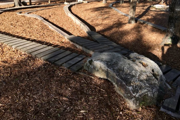 Pathway through a park featuring wooden planks winding around a large rock, surrounded by mulch and shaded by trees.