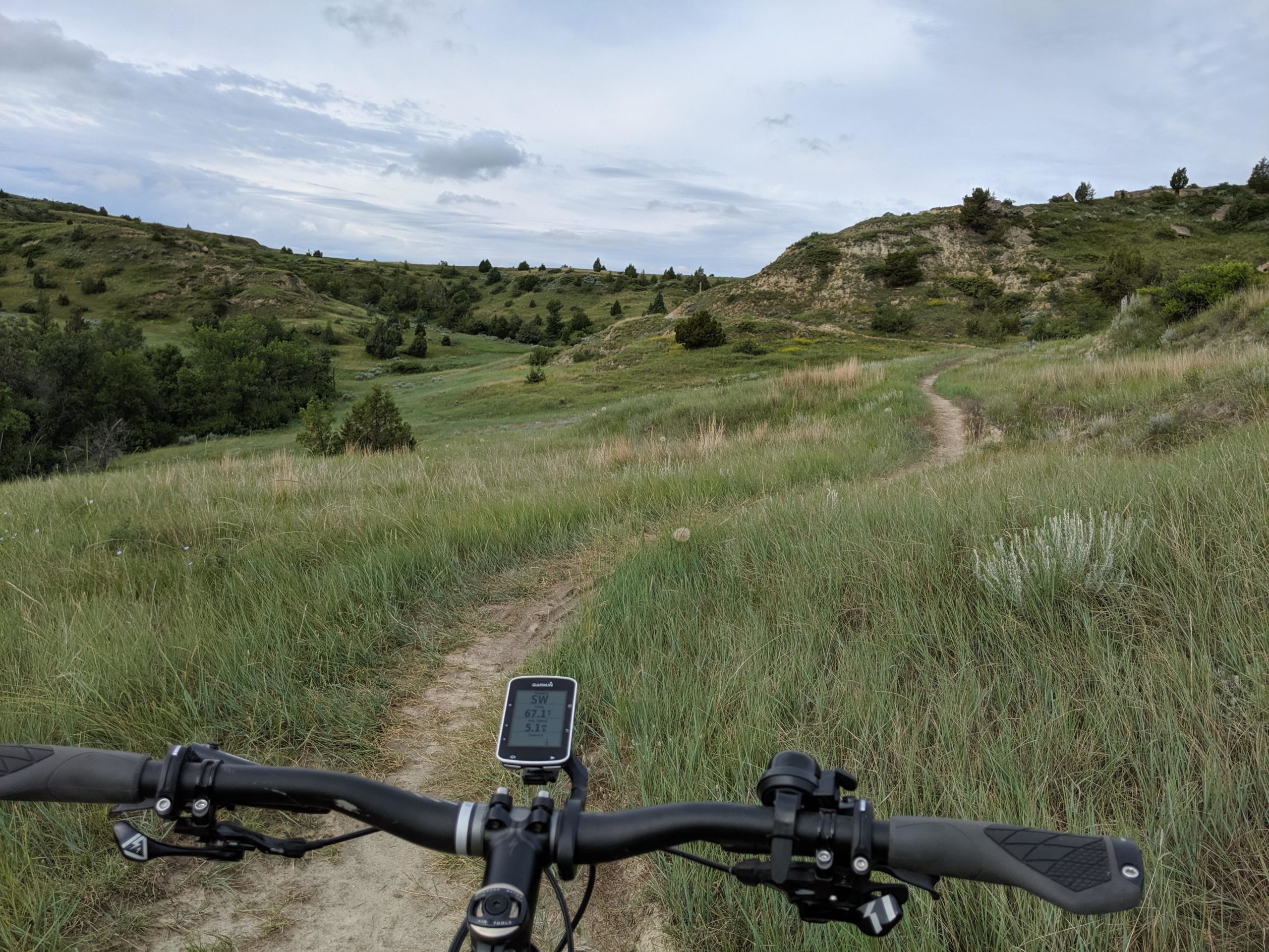 A view from the handlebars of a mountain bike, showcasing a winding dirt path through lush green grass and rolling hills under a cloudy sky. A bike computer displays the current speed and distance metrics. Maah Daah Hey mountain bike trail.