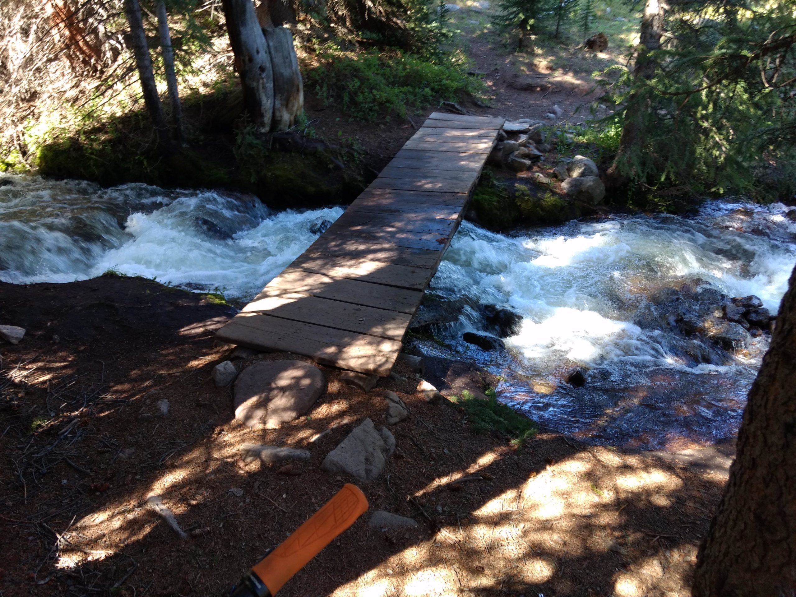 A wooden footbridge spans a rushing stream in a forested area, surrounded by trees and rocky terrain. The water flows energetically beneath the bridge, reflecting light and creating a serene, natural setting. Colorado Trail: Searle Pass and Kokomo Pass (Copper Mountain to Camp Hale) mountain bike trail.