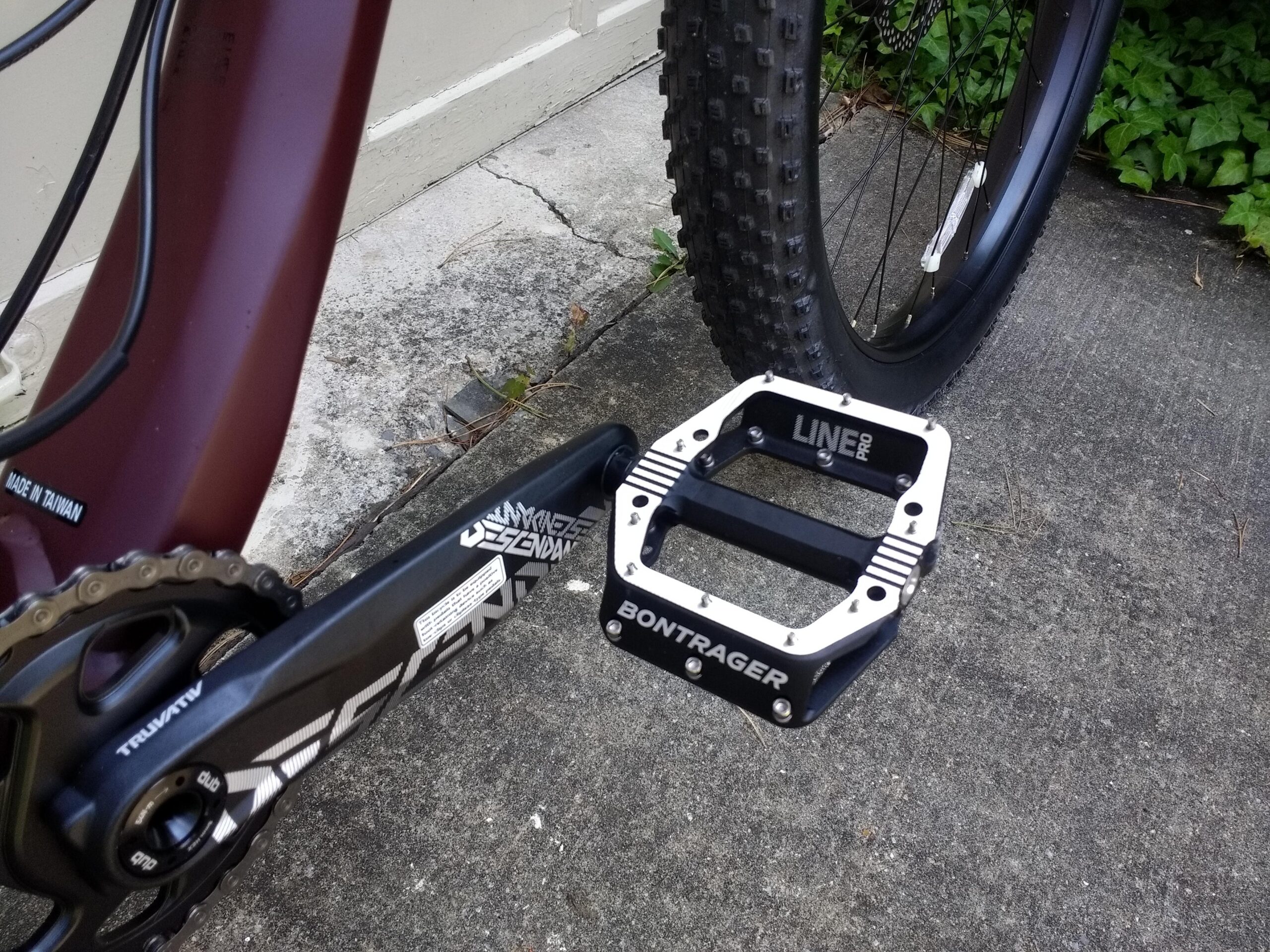 Trek Stache 7: Close-up of a mountain bike's crankset and pedal. The pedal features a silver and black design with the inscription "Bontrager LINE PRO." The bike's frame is maroon, and a portion of the chainring and chain is visible. A textured, knobby tire is partially shown in the background, resting on a concrete surface with some greenery nearby.