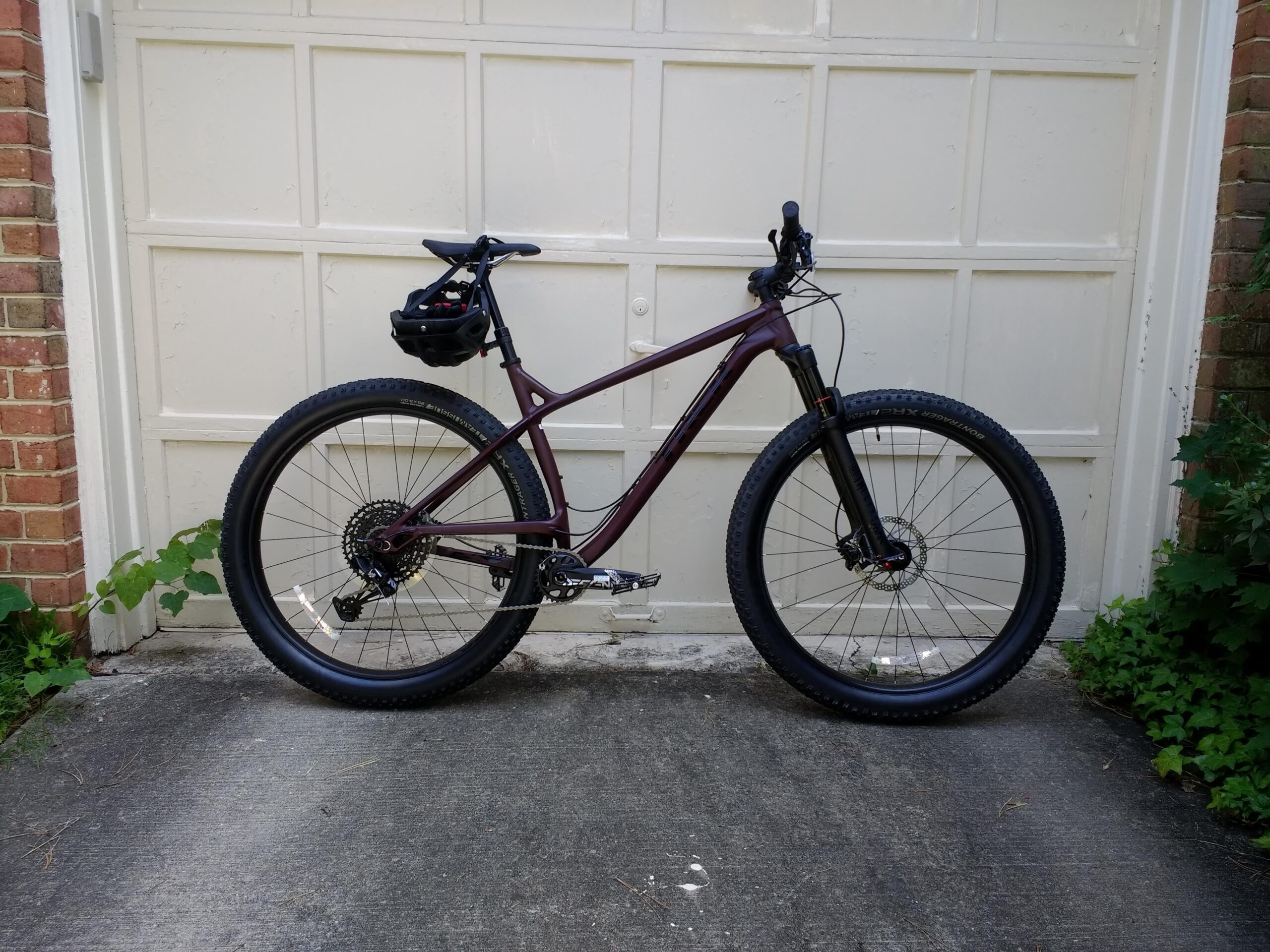 Trek Stache 7: A dark purple mountain bike with wide tires and a gear system, parked on a concrete surface in front of a white garage door. A small backpack is attached to the bike's frame. Green foliage is visible in the background.