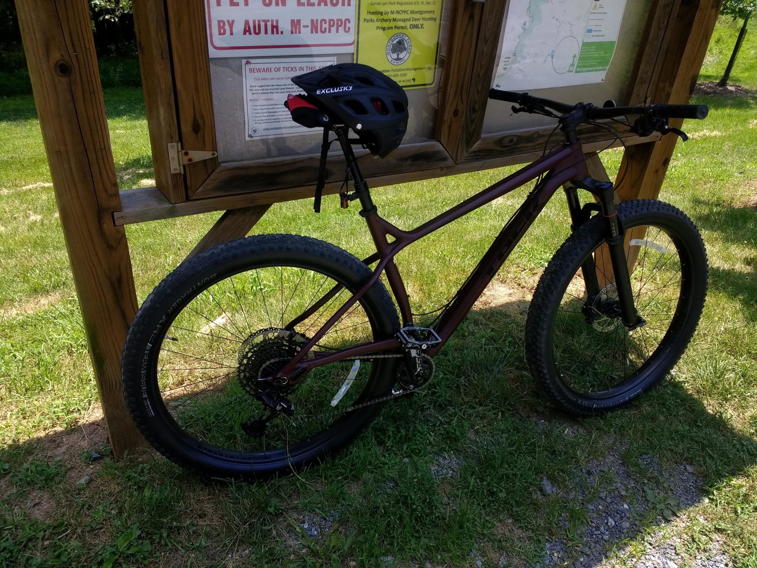 A mountain bike with wide tires is parked next to an information board in a grassy area. The bike is maroon and features a helmet attached to the handlebars. The board displays regulations and a map for visitors. Seneca Creek Greenway Trail mountain bike trail.