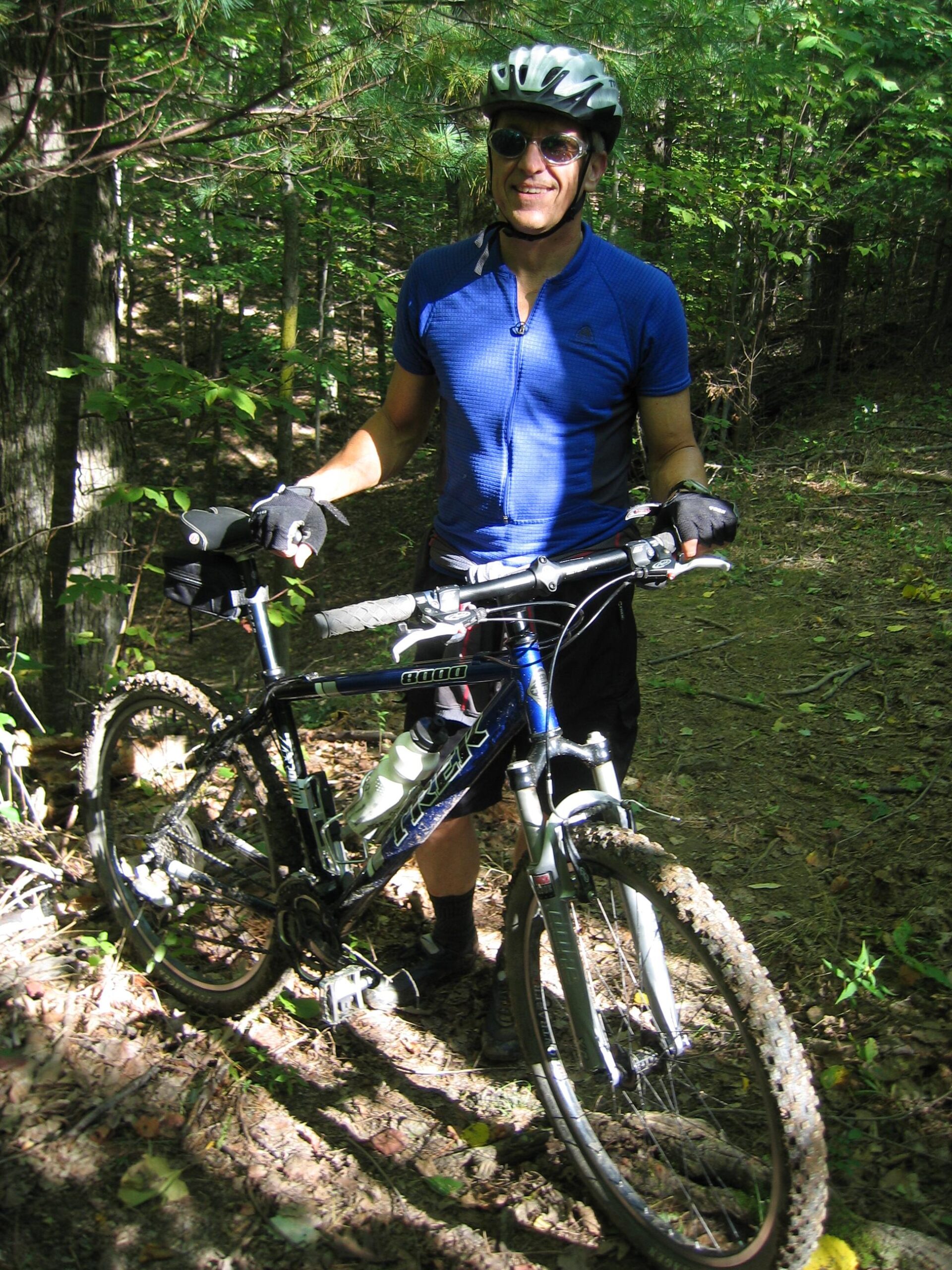 Trek 8000: A person in a blue cycling jersey and helmet stands next to a mountain bike in a forested area, smiling at the camera. The trail is surrounded by green trees and underbrush, with dappled sunlight filtering through the leaves.